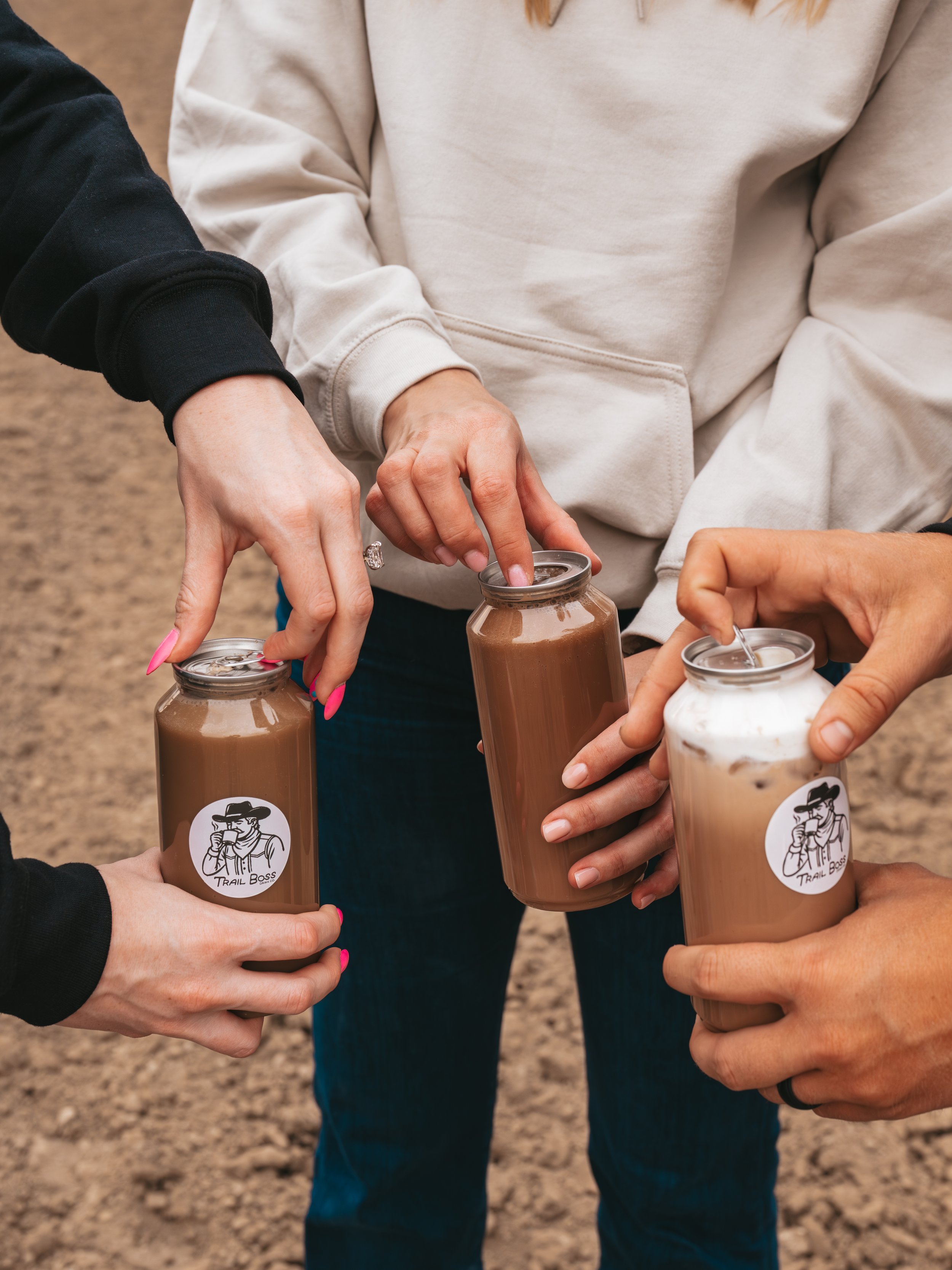 Four people holding plastic cups filled with chocolate milkshake, with visible lids and straws, close-up of their hands and torsos, in an outdoor setting.