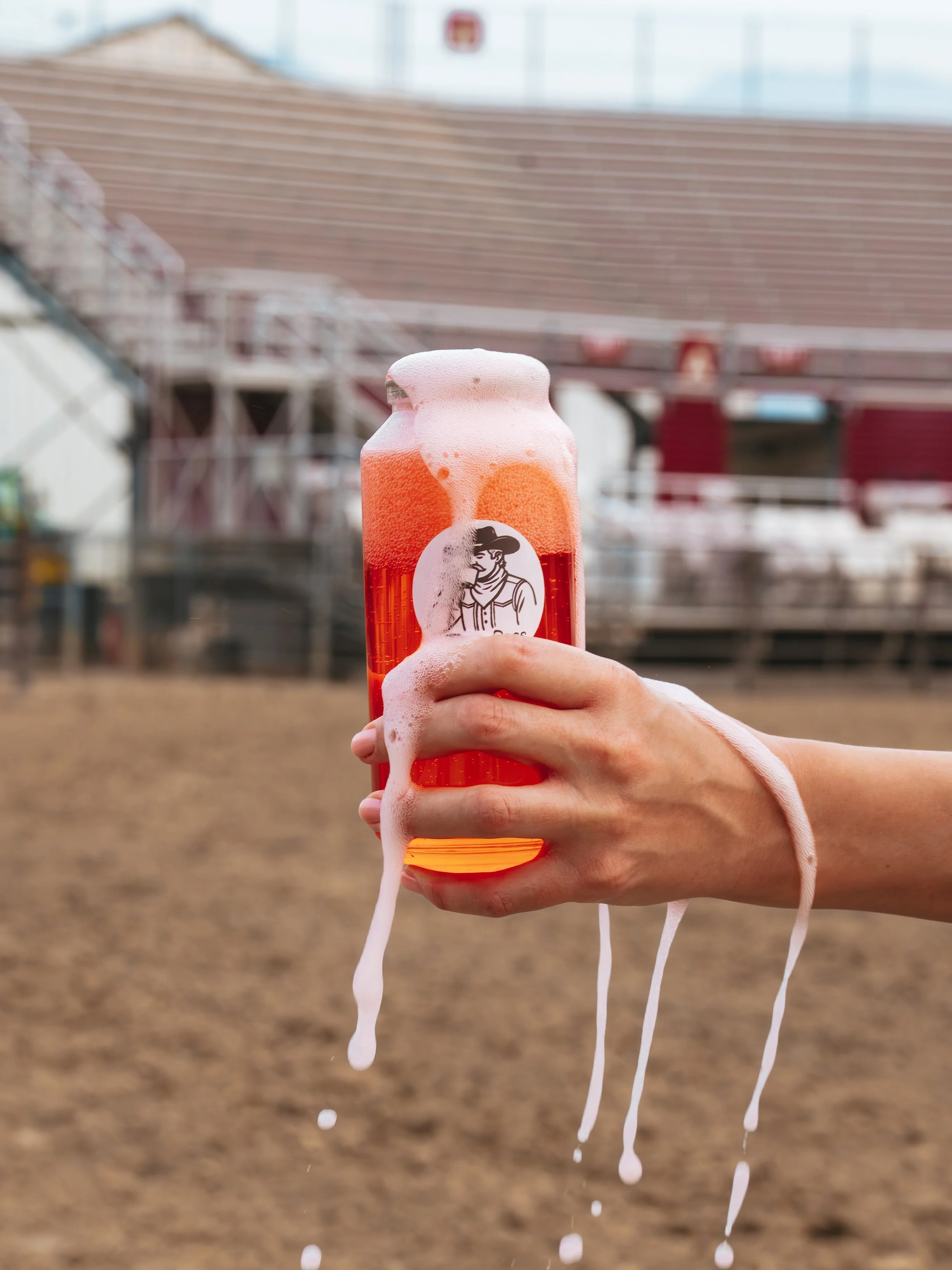 Close-up of a hand holding a red bottle with a foam cap and foam spilling over the sides. The background shows empty bleacher seats in an outdoor stadium.