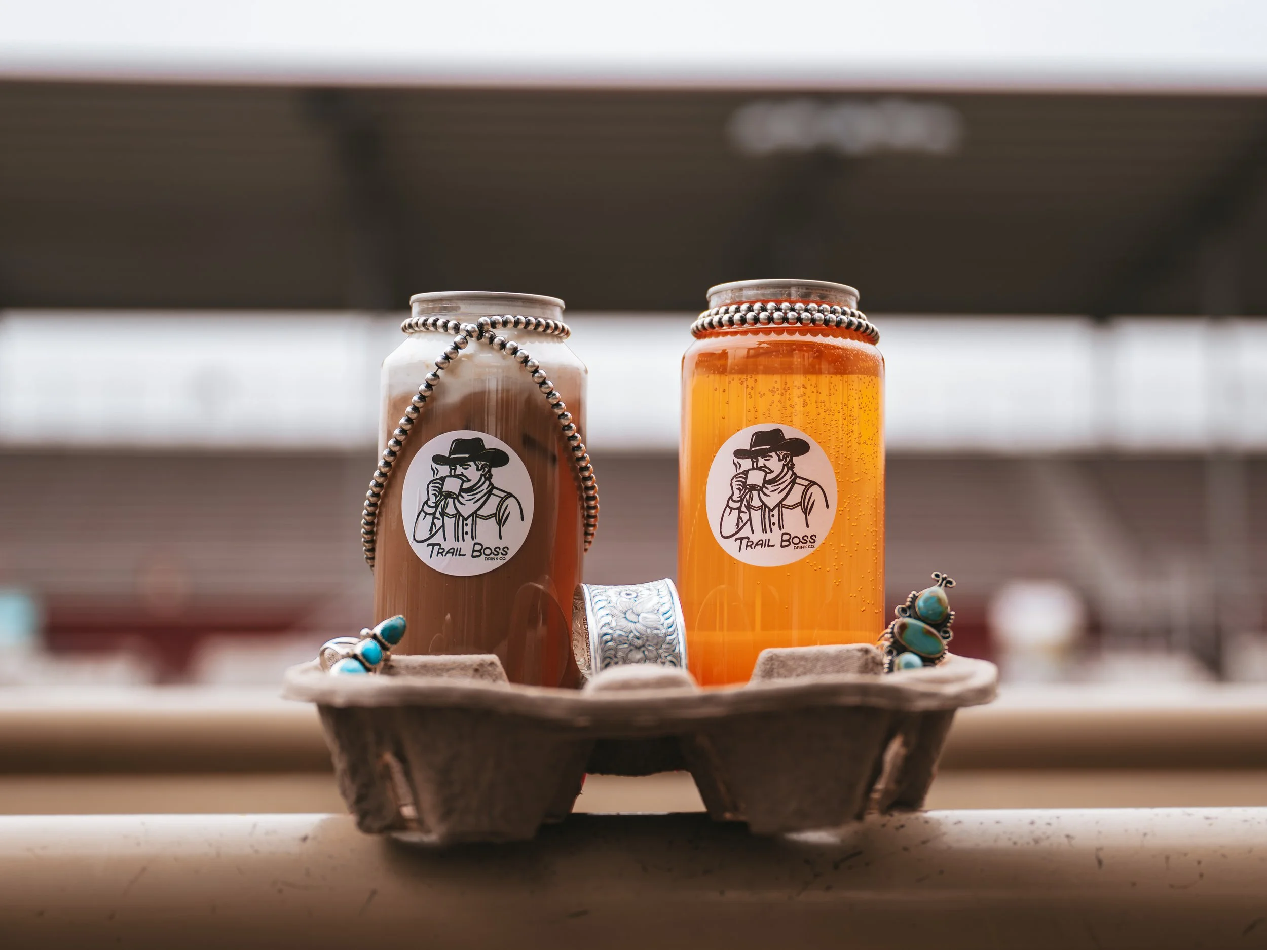 Two bottles of beverage on a tray, one with a white label showing a person drinking from a mug, and the other with an orange liquid, both decorated with beaded necklaces, on a table with jewelry around.
