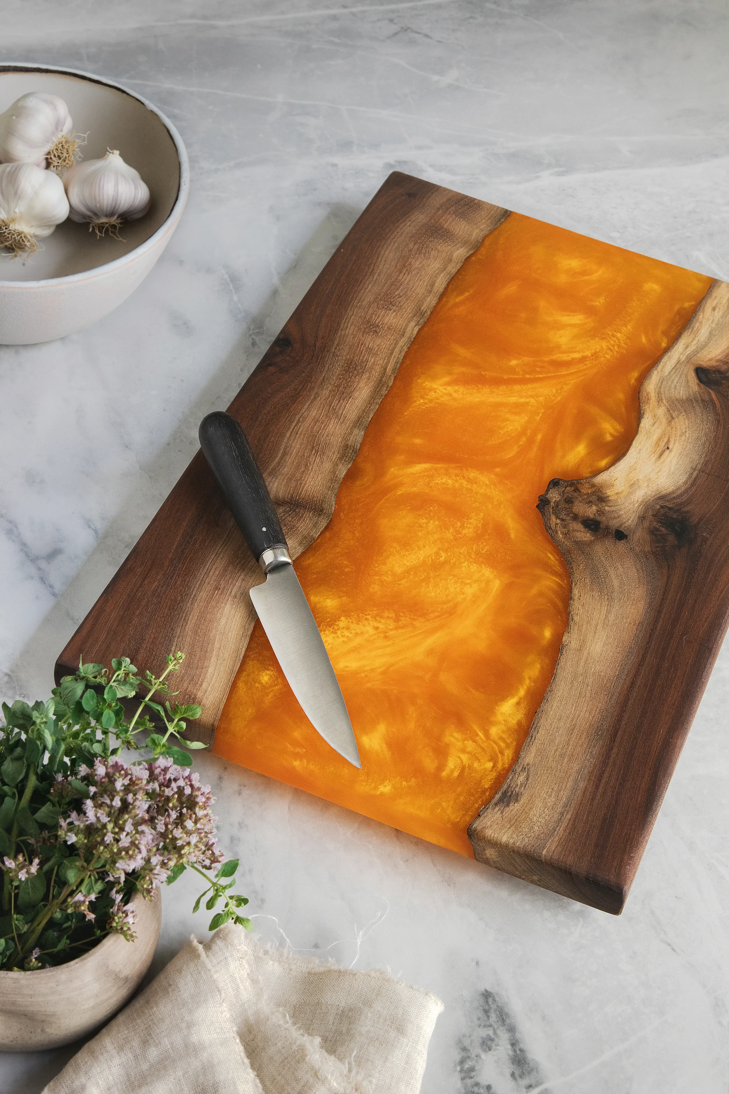 A wooden cutting board with a slice of butternut squash, a knife, a potted plant with purple flowers, and a bowl of garlic cloves on a light marble surface.