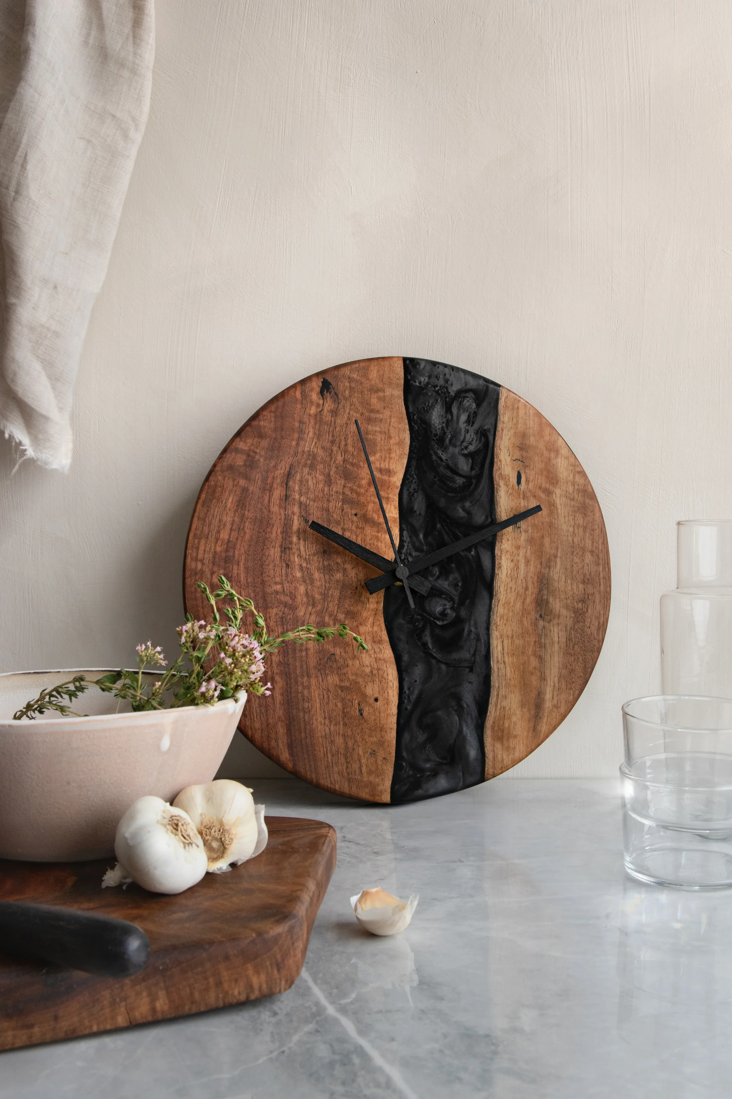 Close-up of a round wooden wall clock with black and carved face, on a kitchen counter next to a bowl of flowers, garlic, and a glass of water.