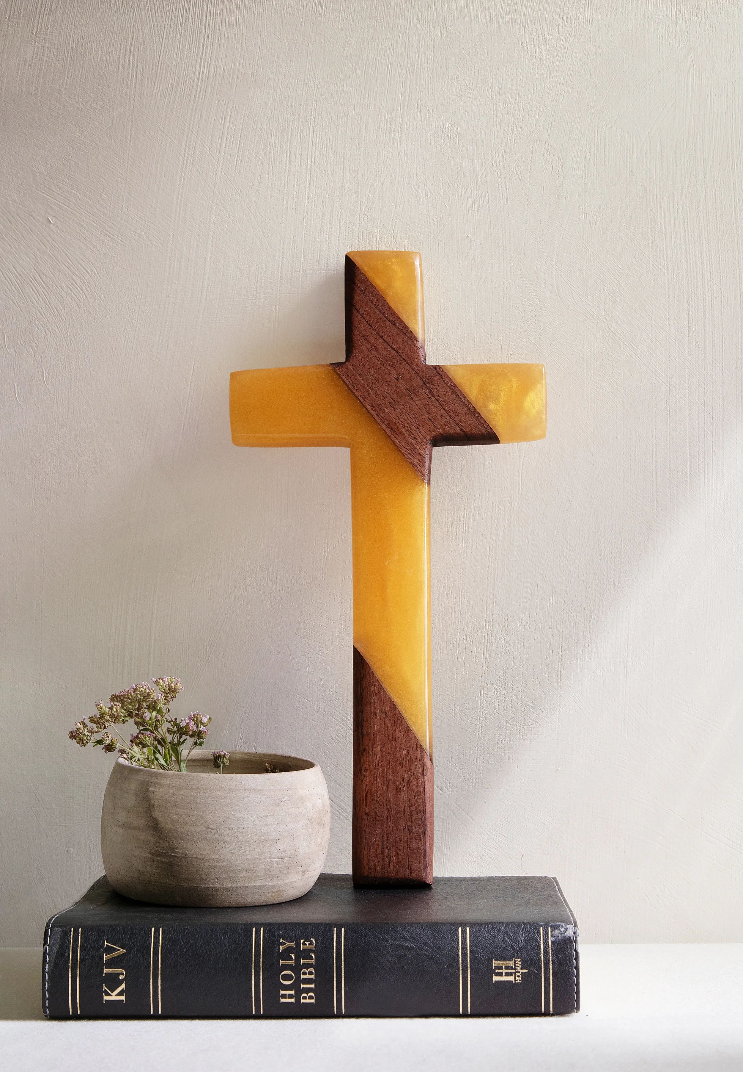 A decorative religious display with a large cross made of wood and yellow material, placed on top of a black Holy Bible, and a small white pot with flowers on the left side, all against a plain wall background.