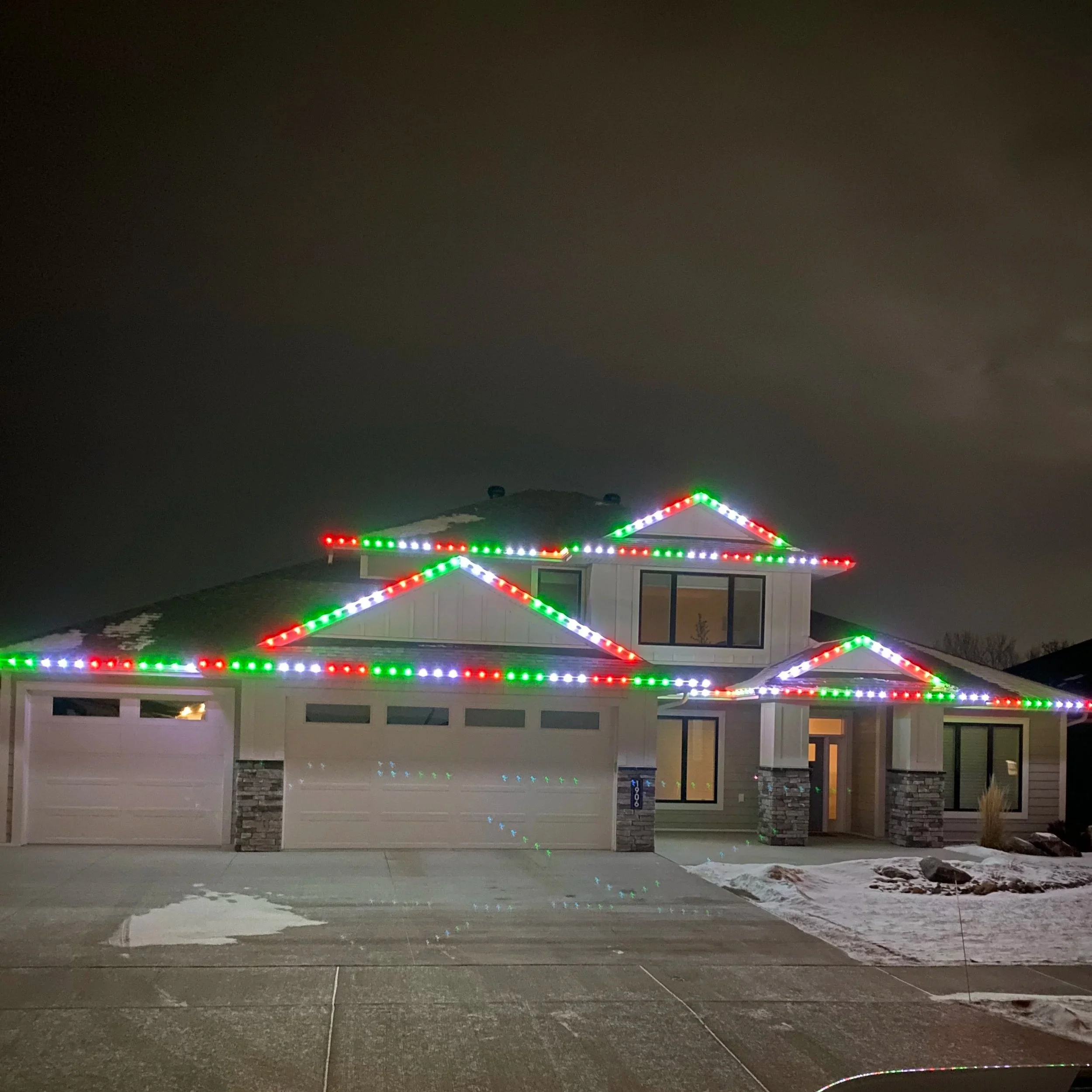 A house decorated with colorful Christmas lights on its roofline and roof edges, taken at night with a dark sky and some snow on the ground in front.
