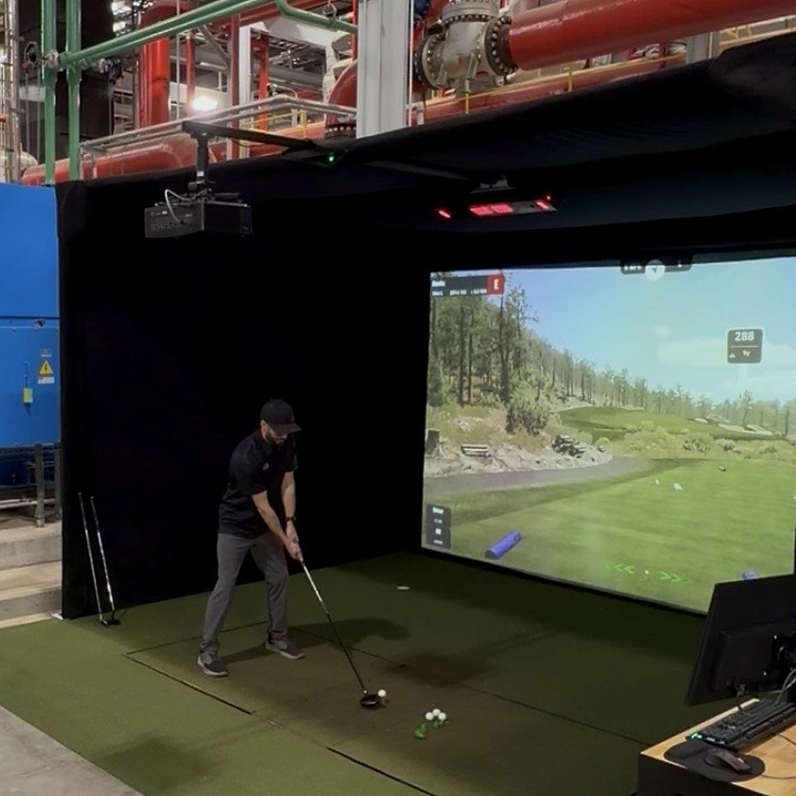 Person playing virtual golf on a simulated golf course indoors, with a large screen displaying a golf landscape and realistic visuals.