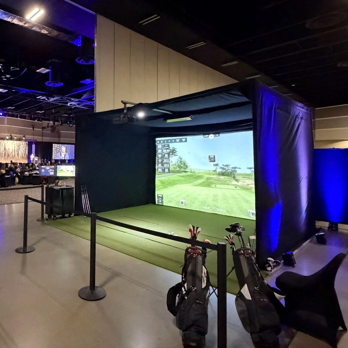 Indoor golf simulation setup with a large screen showing a golf course, golf clubs and bags, and a small seating area with a black barrier.