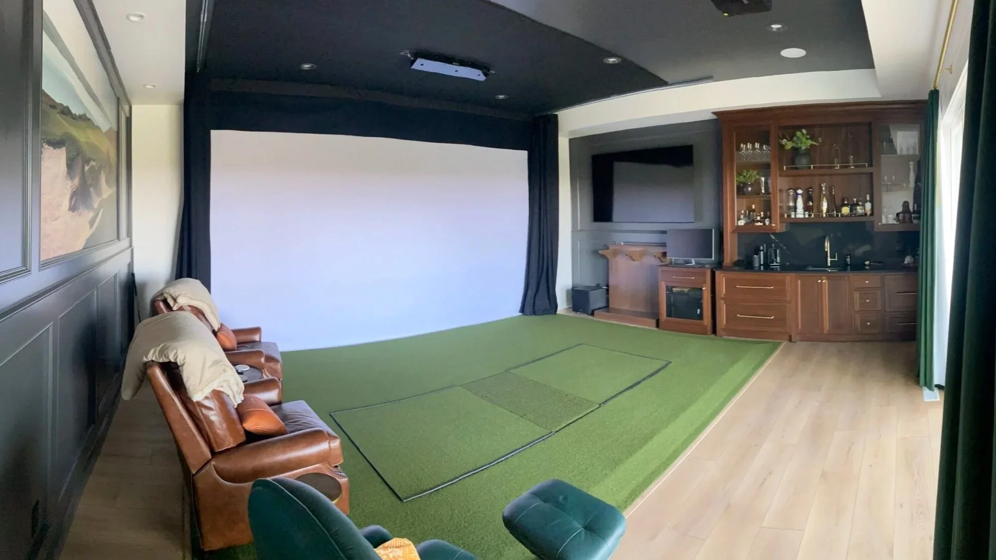 A room with a large white projection screen on the wall, a green carpeted floor with some sections outlined, a small bar with shelves containing bottles, a television on a wooden stand, a few leather chairs along the wall, and a window with green curtains.