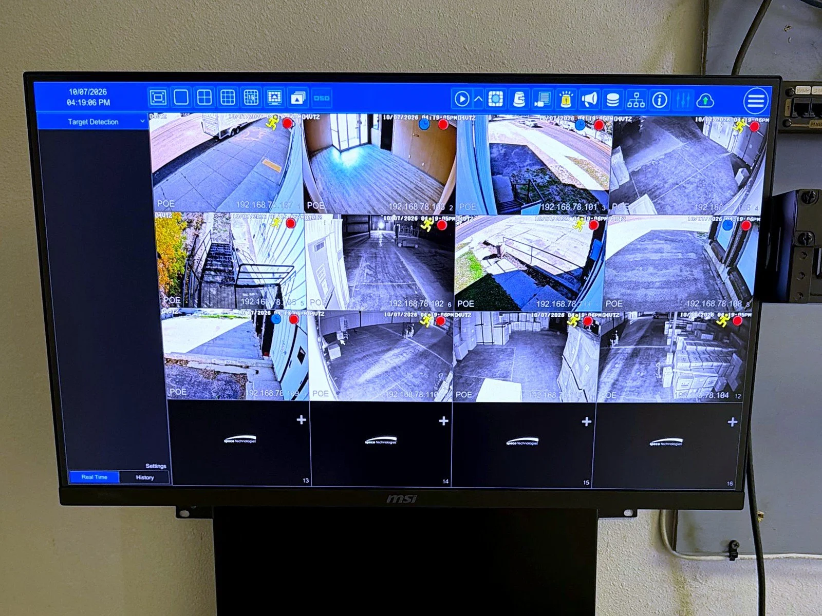 Security camera monitor displaying multiple live footage feeds from different outdoor and indoor locations, showing areas such as a parking lot, building entrance, alleyway, and storage area.