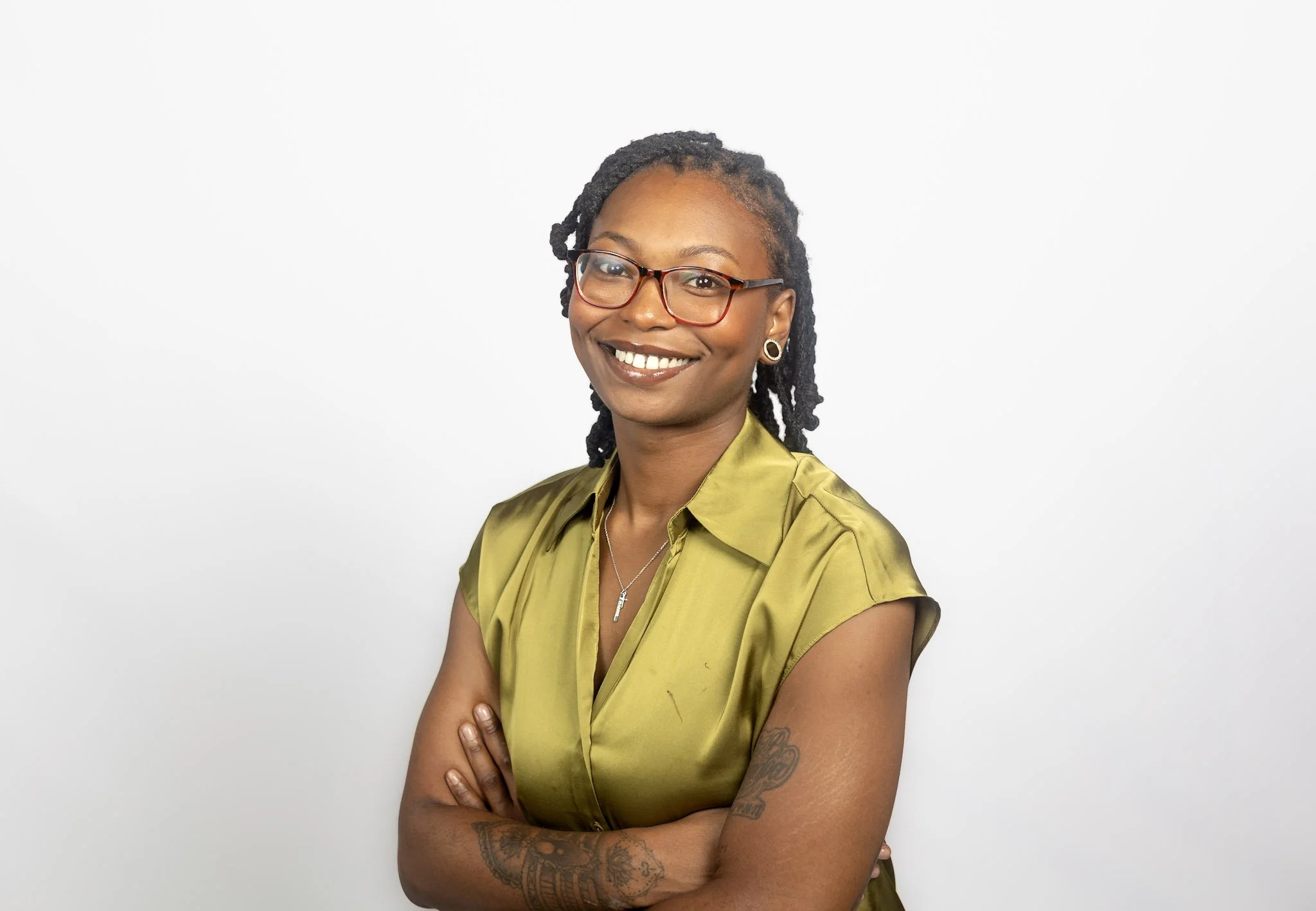 A woman with glasses, wearing a green satin shirt, smiling, with arms crossed, standing against a white background.