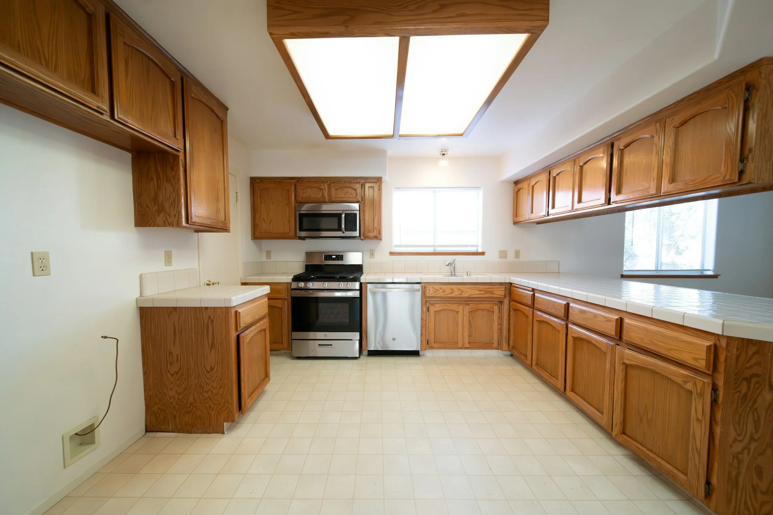 Kitchen with wooden cabinets, white countertop, stainless steel stove, microwave, dishwasher, and a window with blinds.