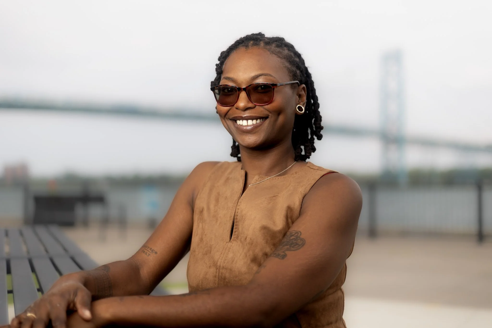 A woman with dark curly hair wearing sunglasses and a sleeveless tan top, smiling, sitting outdoors near a body of water with a blurred bridge in the background.