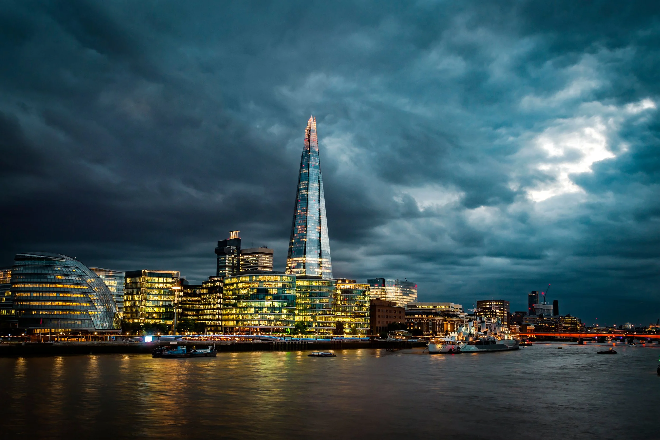 The image shows a city skyline with modern buildings under dark, stormy clouds. The tallest building is a glass skyscraper illuminated with pink and blue lights, and there are boats on a river in the foreground.