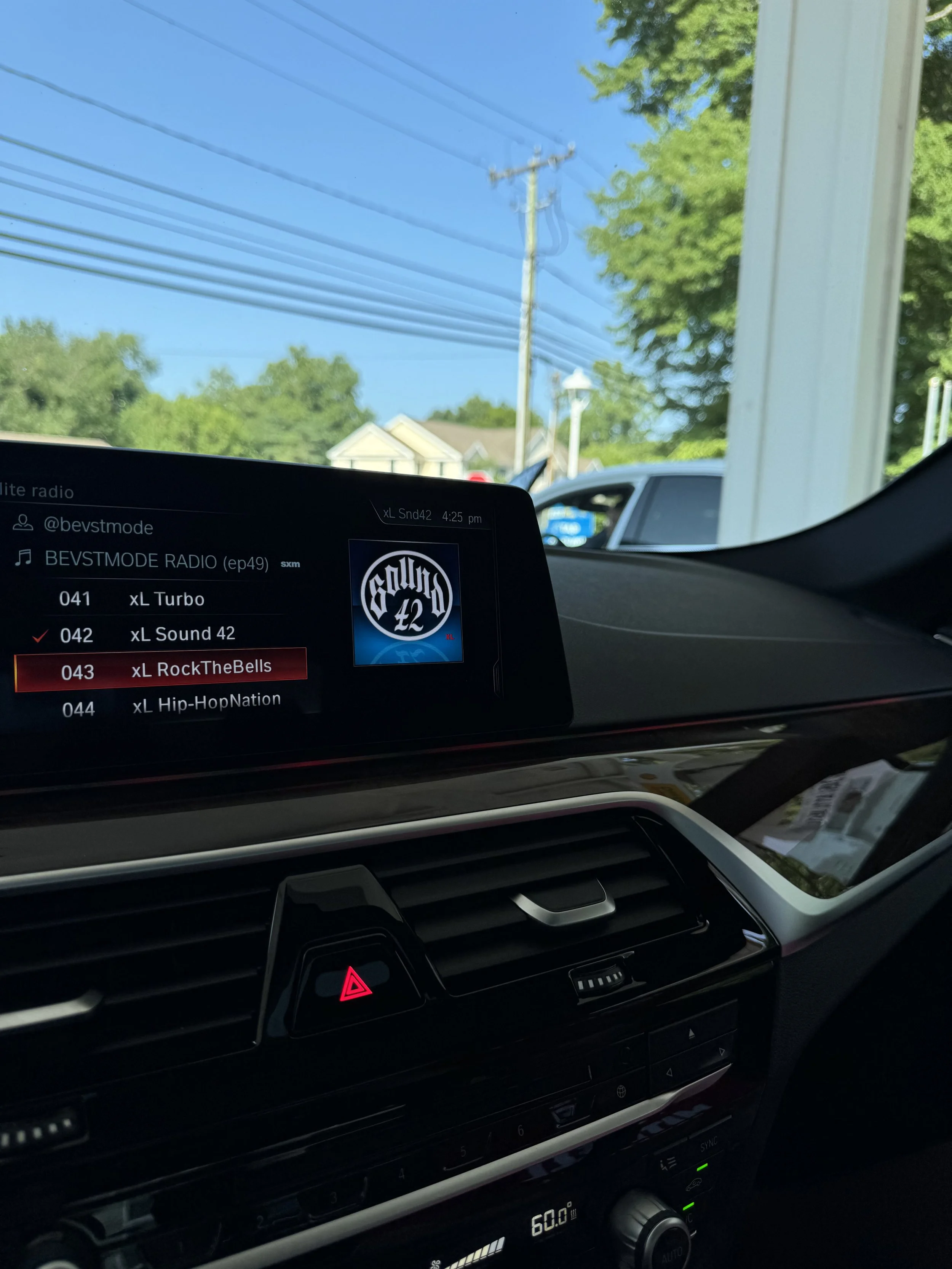 The photo shows the interior of a car with a touchscreen displaying a radio station, with the outside view of a sunny suburban neighborhood with green trees, houses, power lines, and a parked vehicle visible through the window.