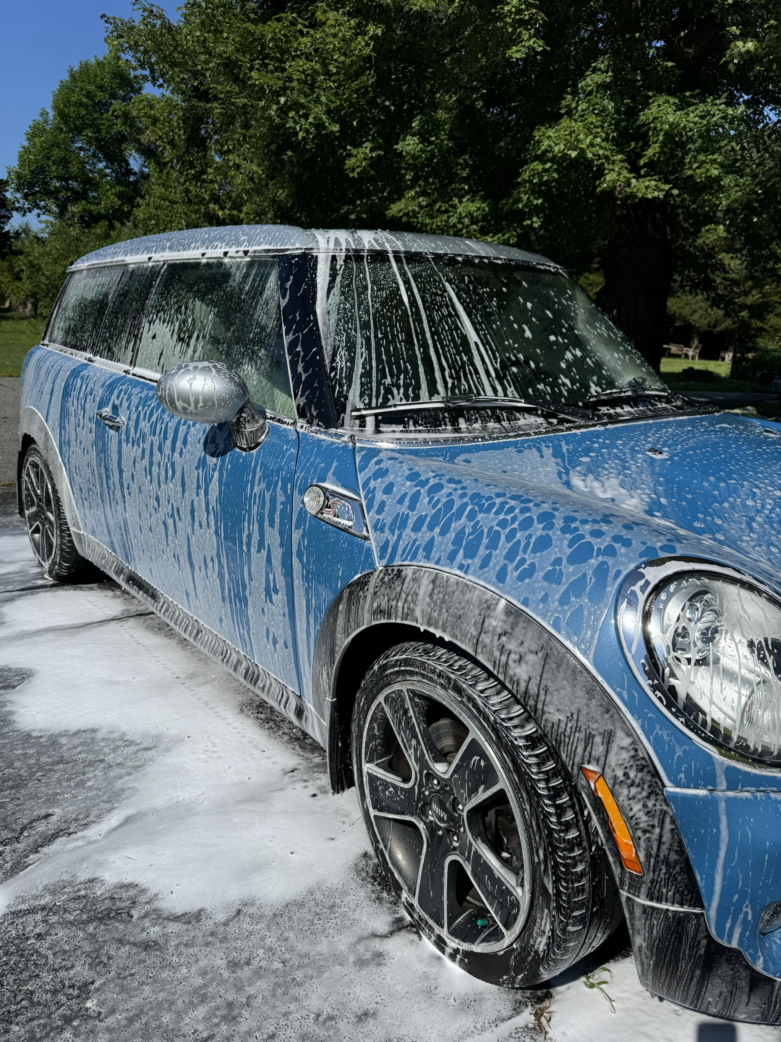 A blue car covered in soap suds after being washed, parked on a driveway with a grassy area and trees in the background.