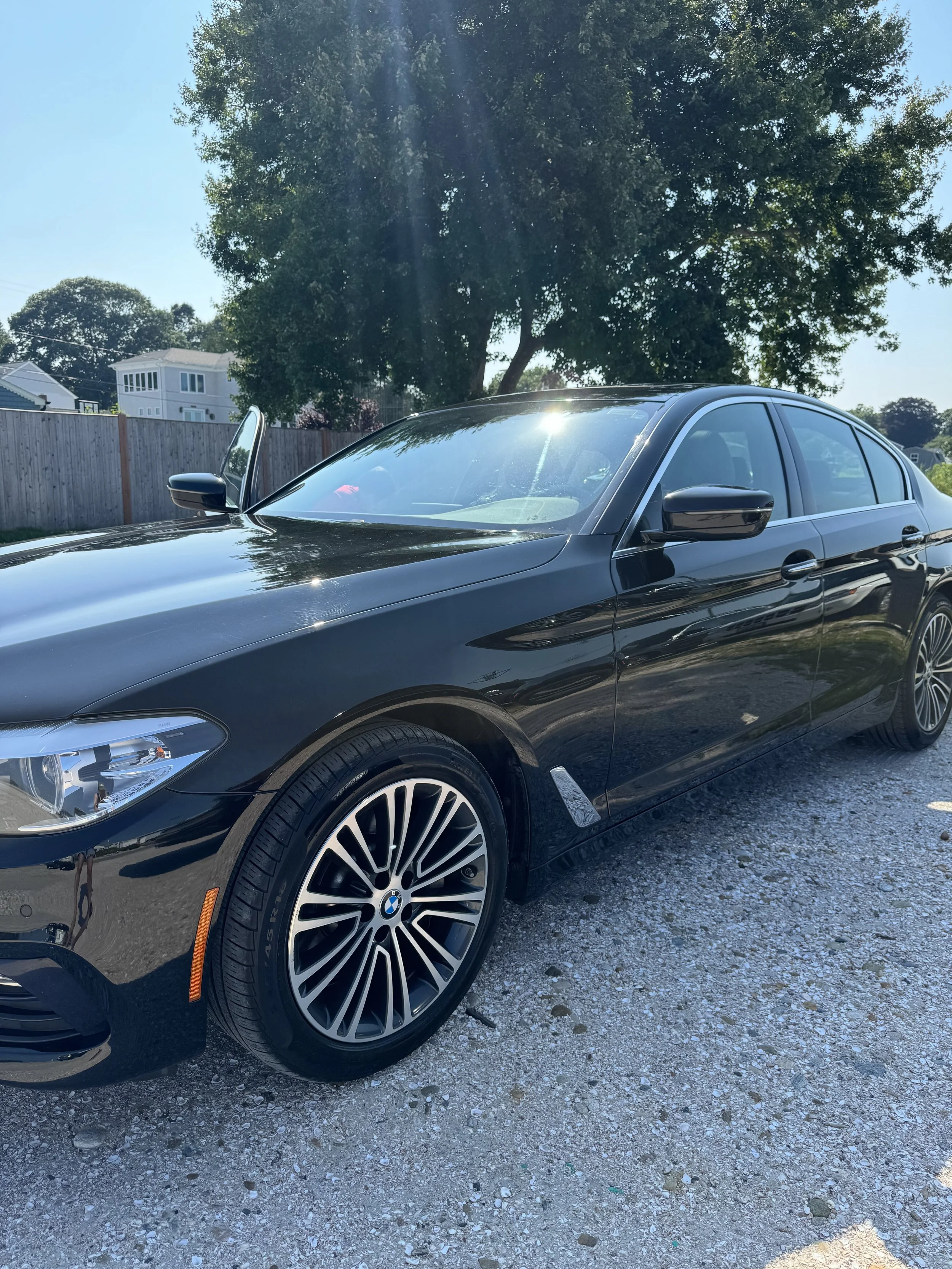 A black BMW sedan parked on a gravel surface with trees and houses in the background and sunlight reflecting off the hood and windshield.