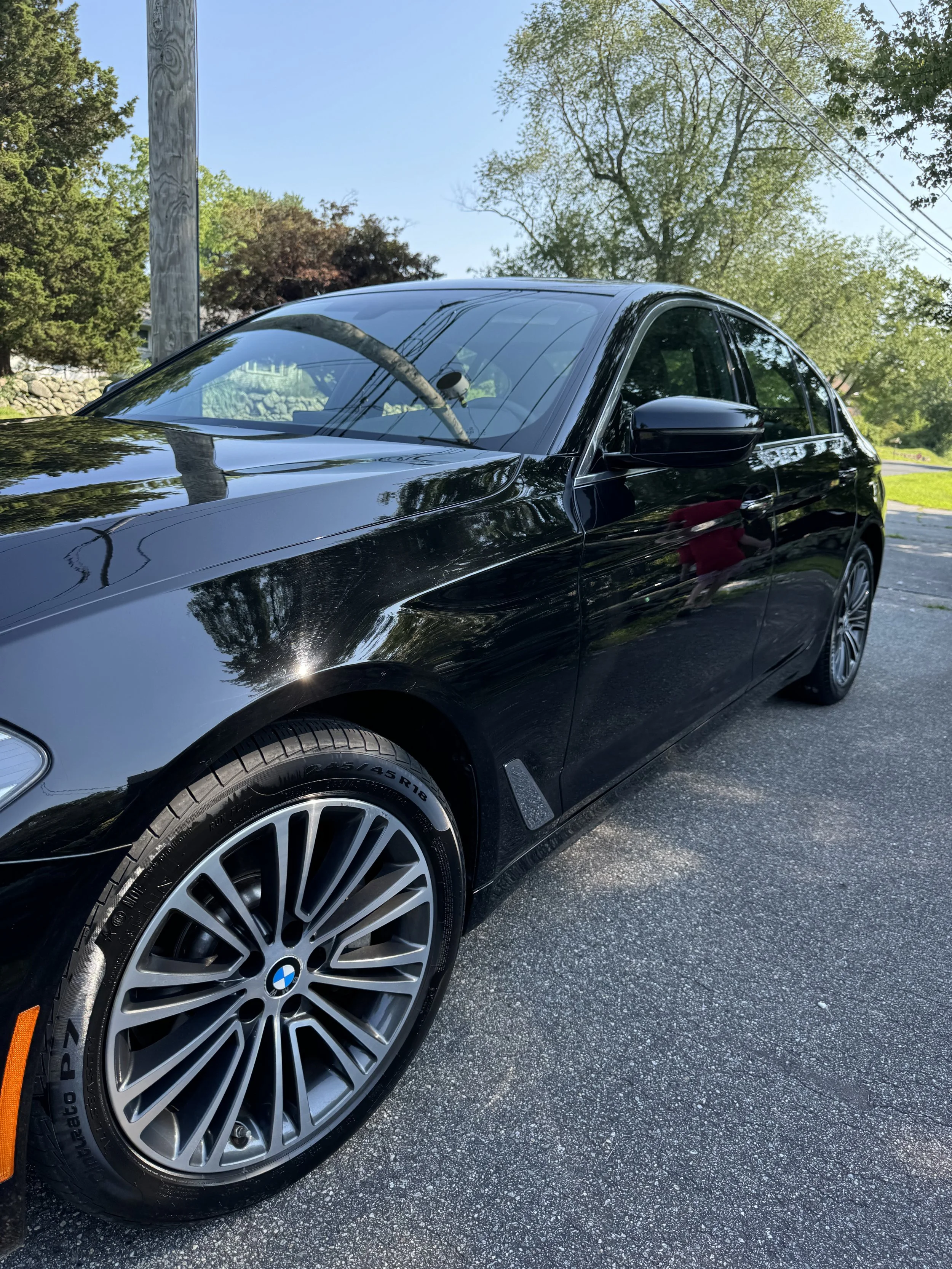 Black BMW sedan parked on a street with trees and power lines in the background, reflecting the surroundings on its shiny surface.