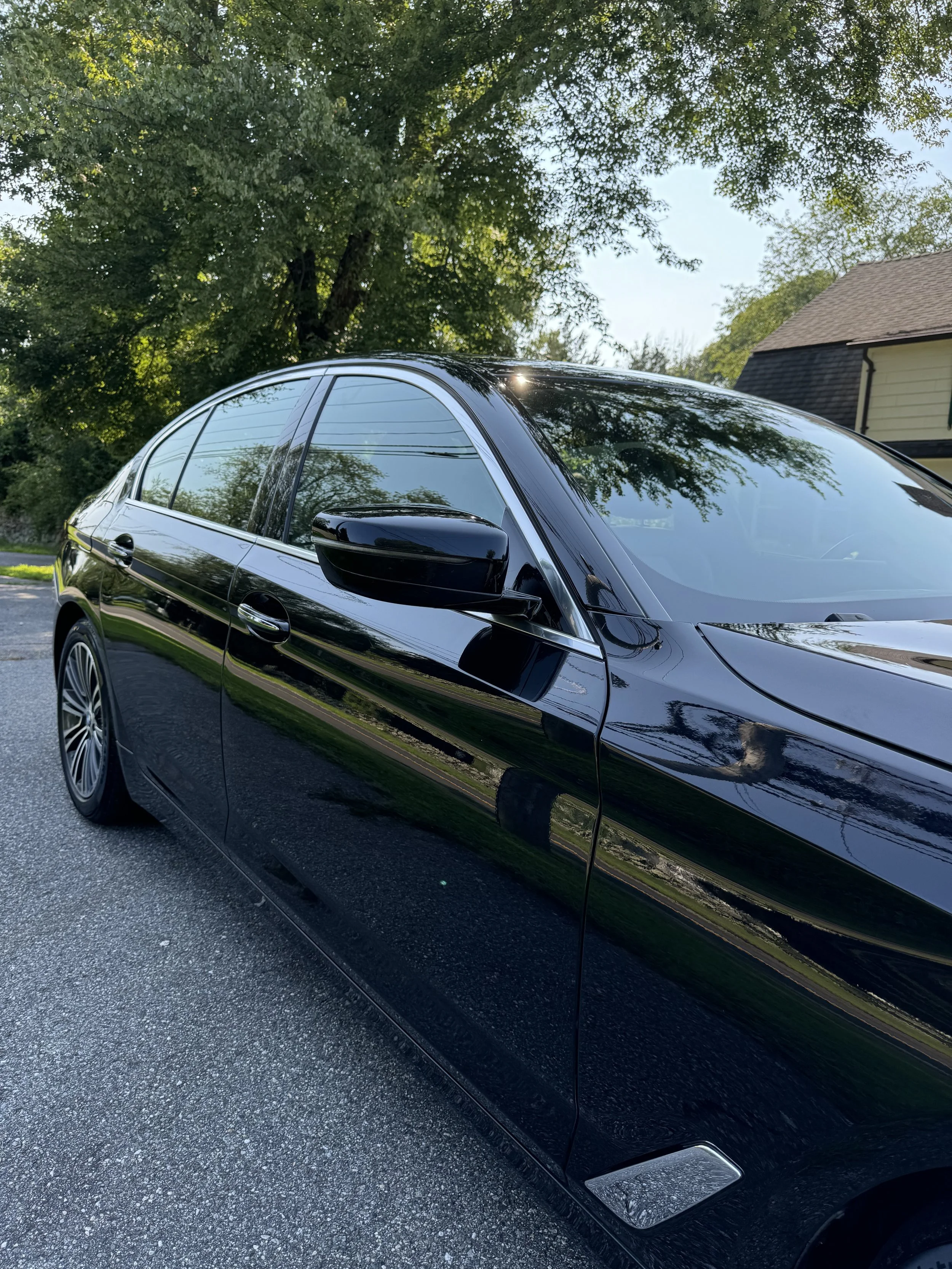 Close-up of a shiny black luxury car parked on a paved street, with trees and a house in the background, and sunlight reflecting off the car's surface.