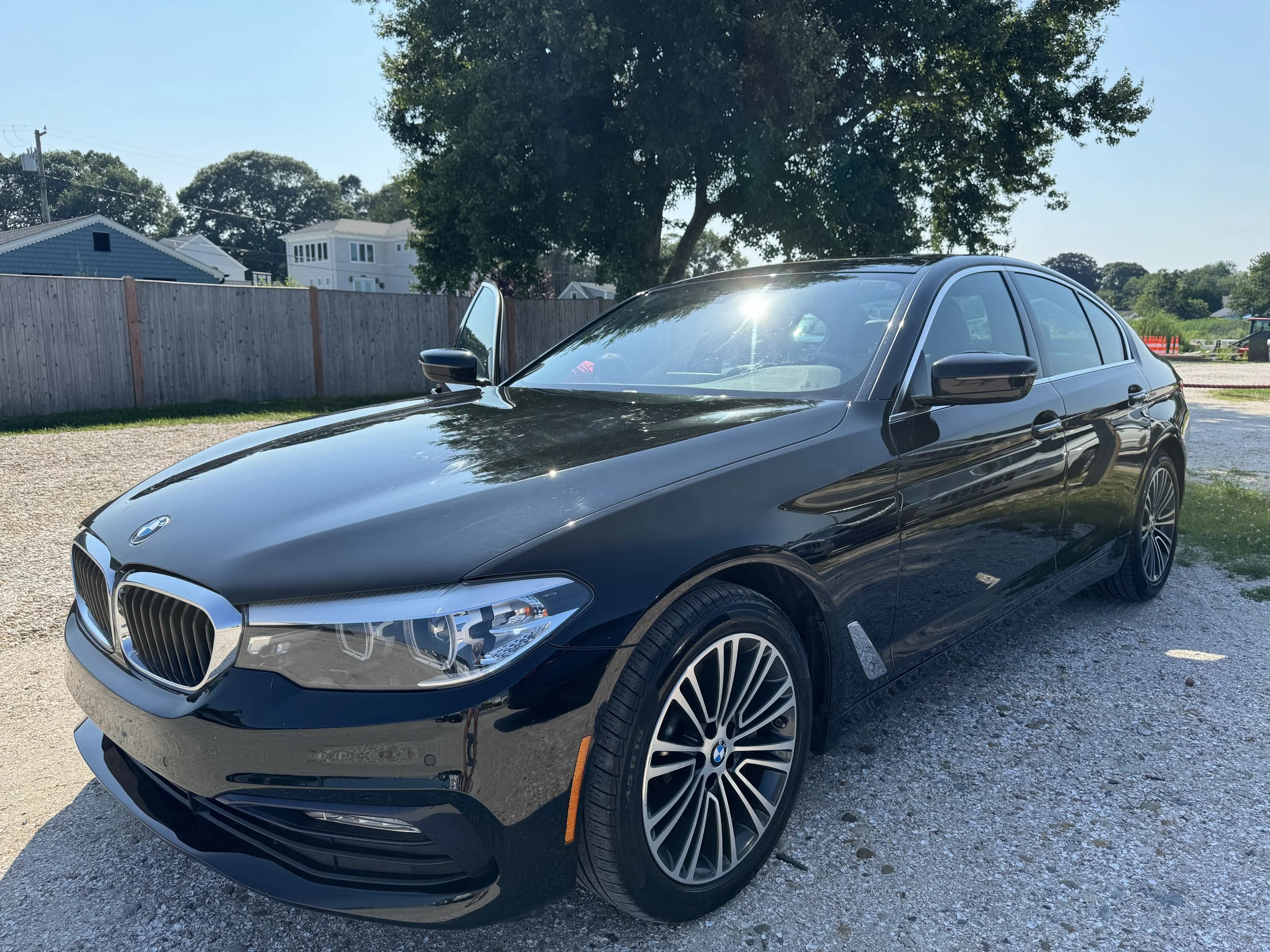 Black BMW car parked on a gravel driveway next to a wooden fence with houses and trees in the background under clear blue sky.