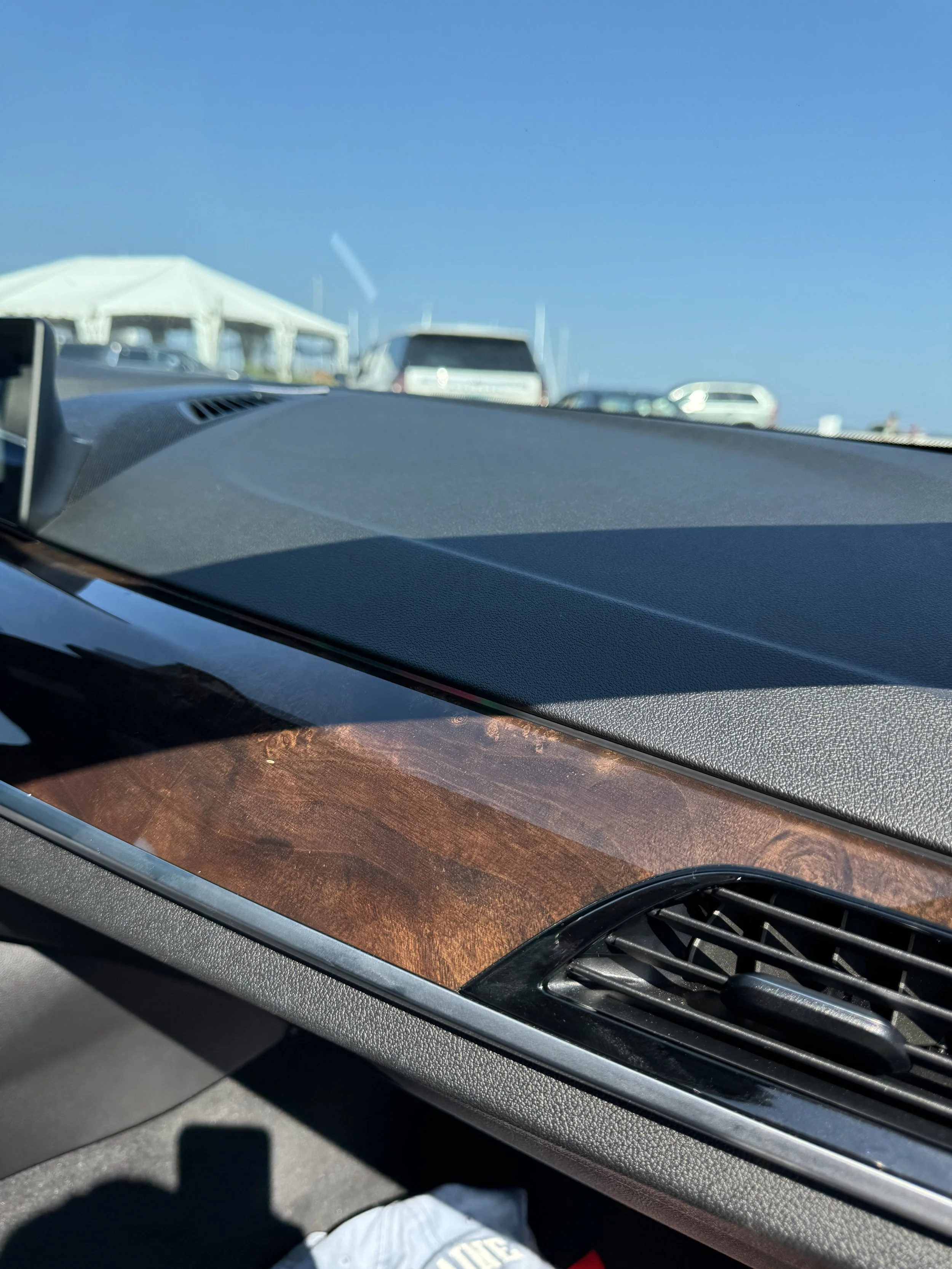 Inside a car windshield showing dashboard with wood panel, air vent, and part of a person's white clothing and shoe at the bottom.