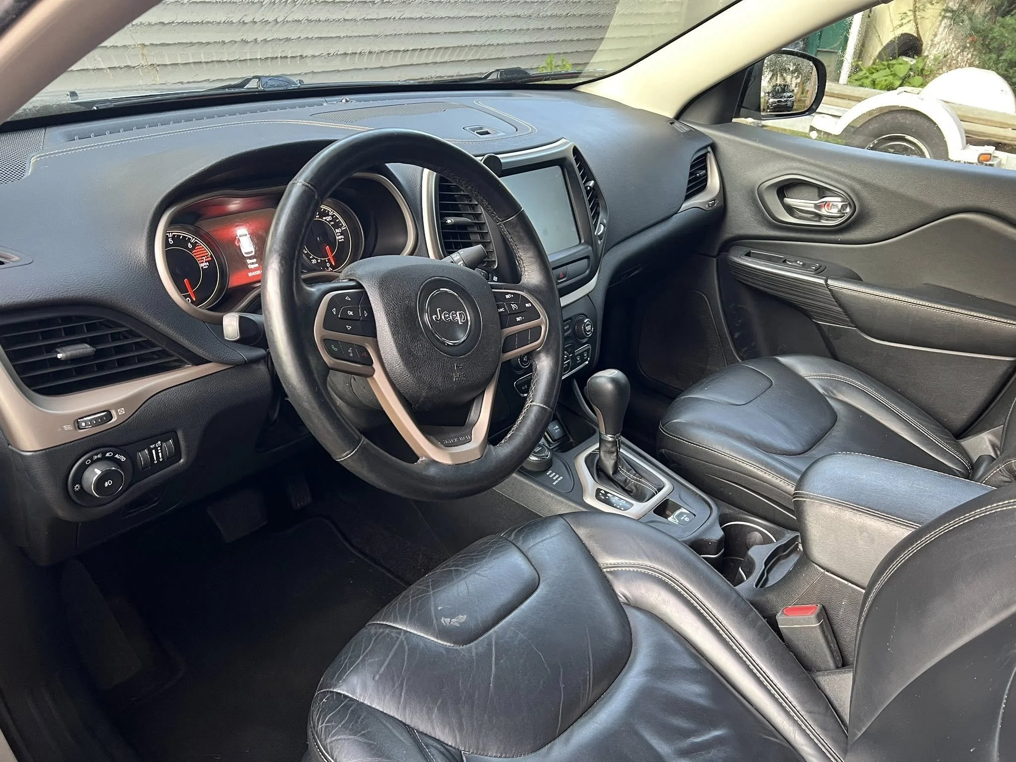 The interior of a Jeep vehicle showing the steering wheel, dashboard with speedometer and tachometer, gear shift, and black leather seats. Outside the vehicle, part of a boat and a golf cart are visible through the passenger side window.