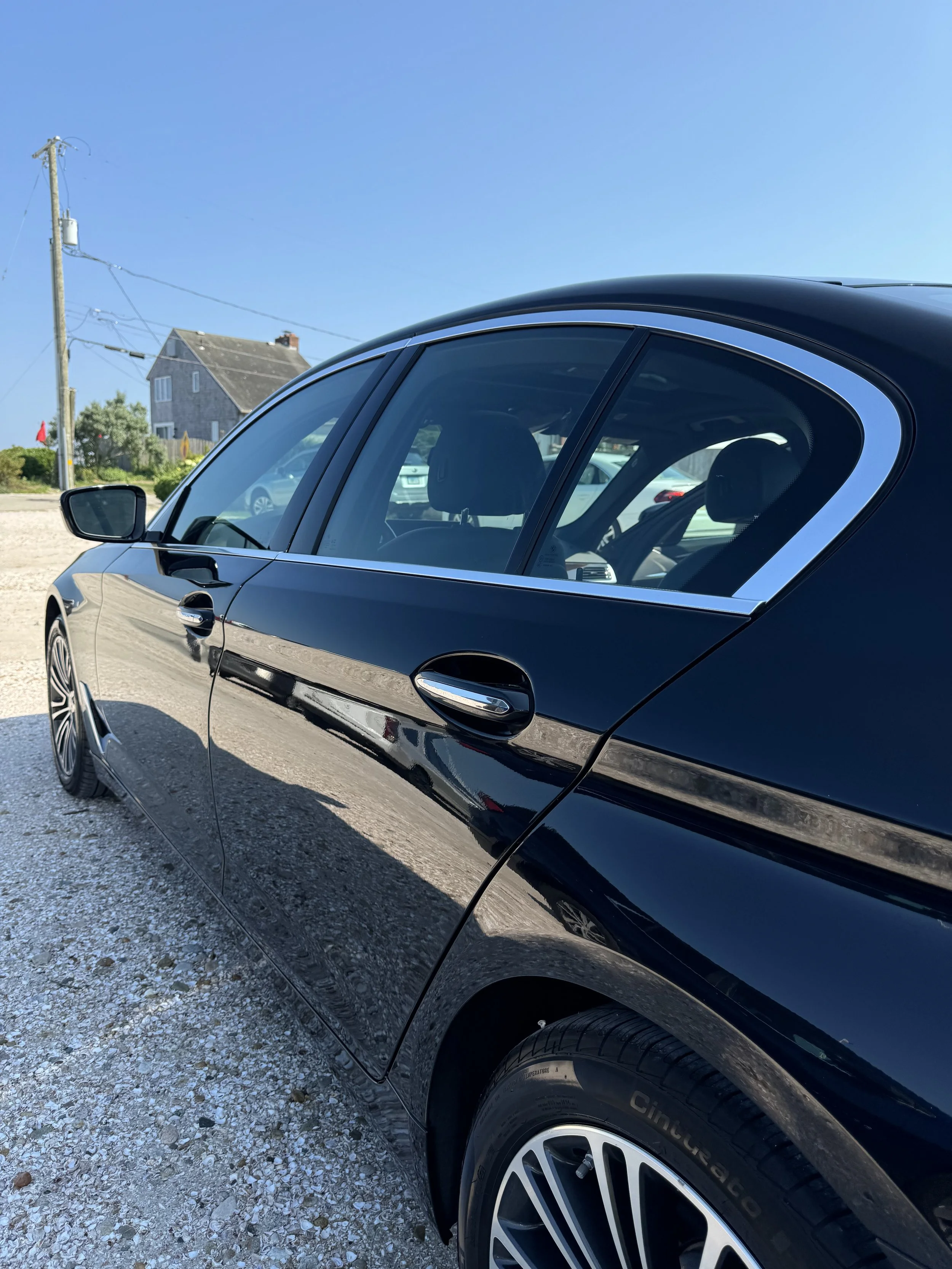 A black luxury car parked outdoors on a gravel surface during daytime. In the background, there is a house, some greenery, and a utility pole with power lines under a clear blue sky.