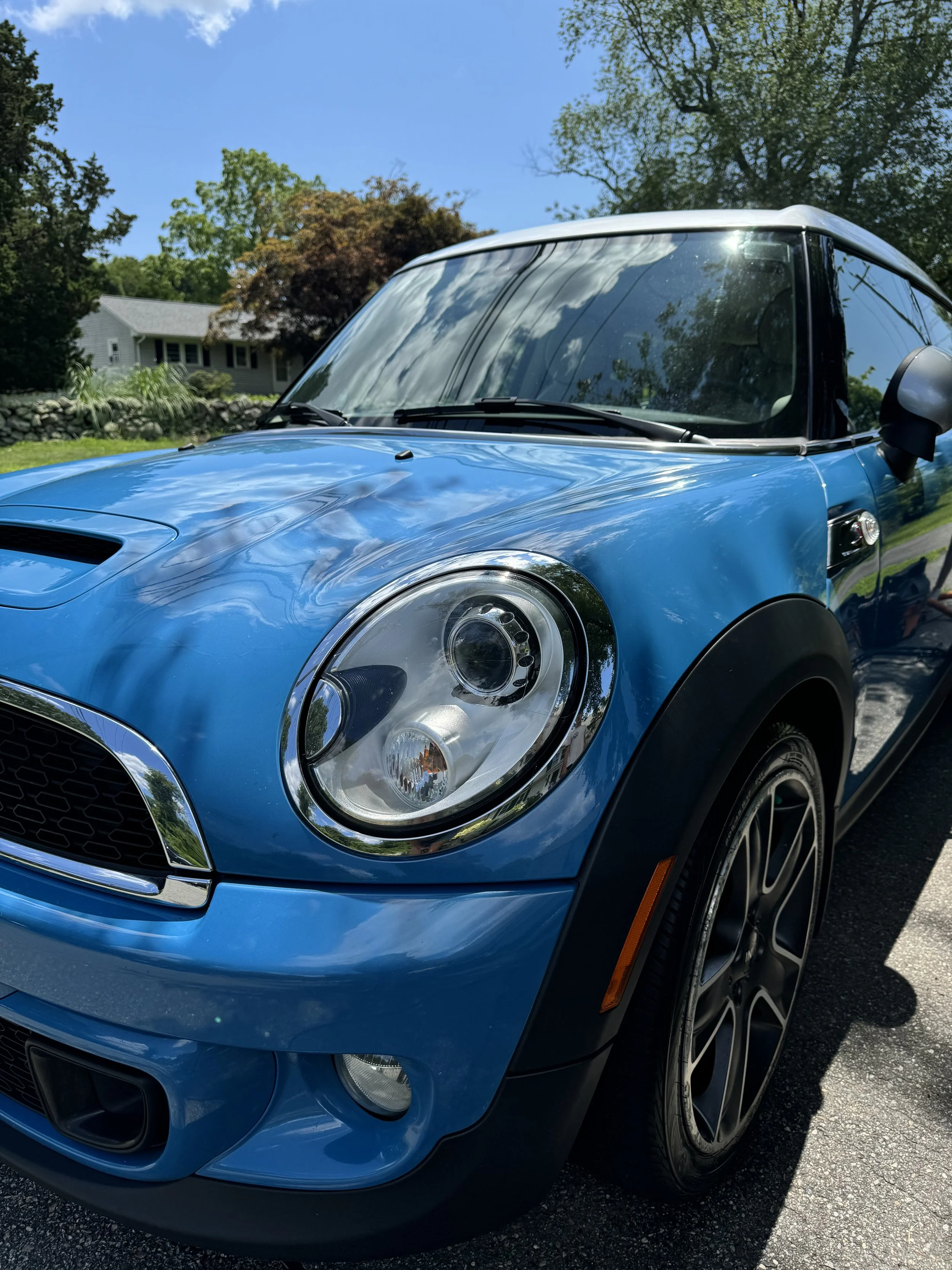 Close-up of a blue Mini Cooper parked outdoors on a sunny day, with reflection of trees and sky on the windshield.