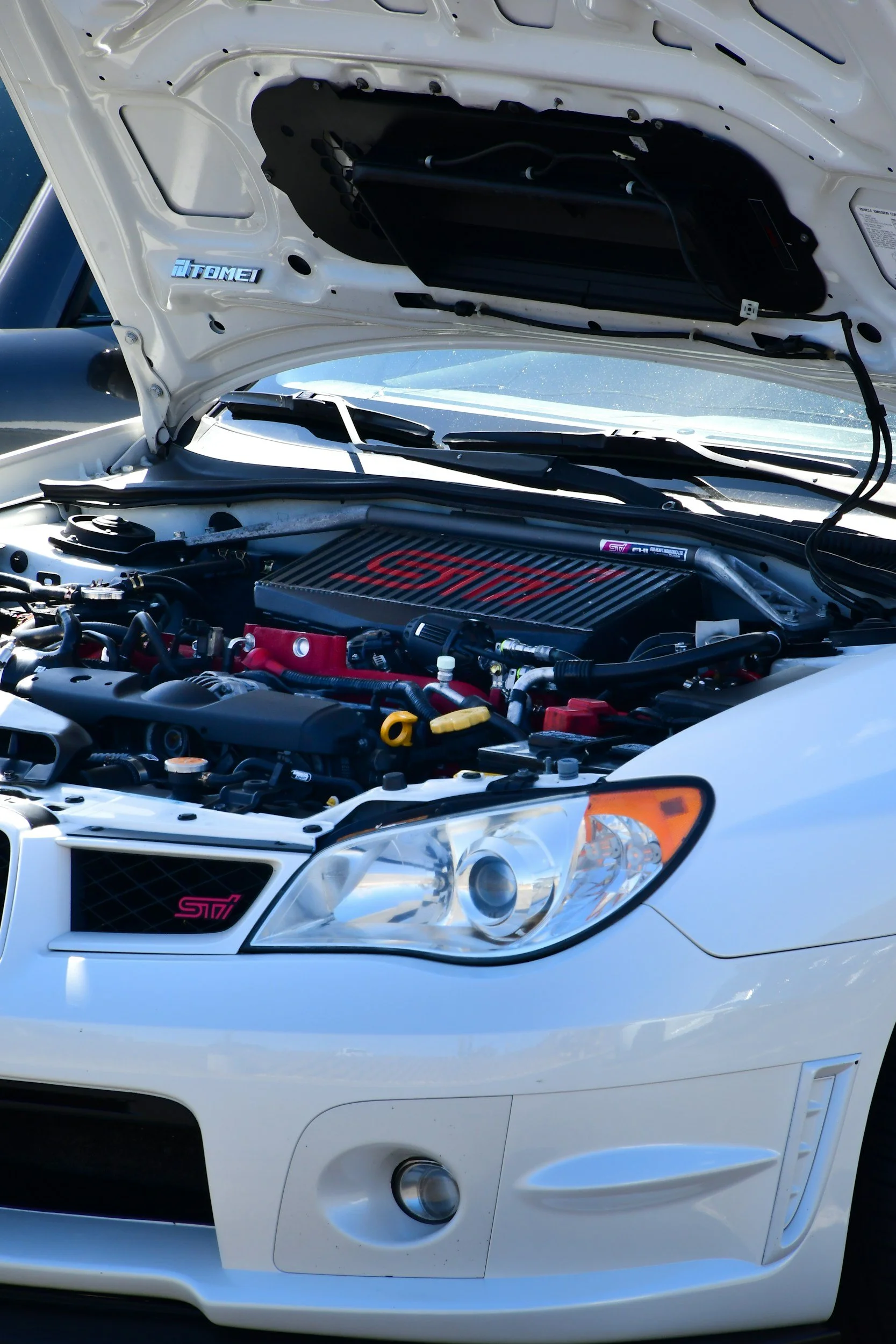 Front view of a white Subaru WRX STI sports car with its hood open, revealing the engine. The car has a pink STI badge and a black front grille.
