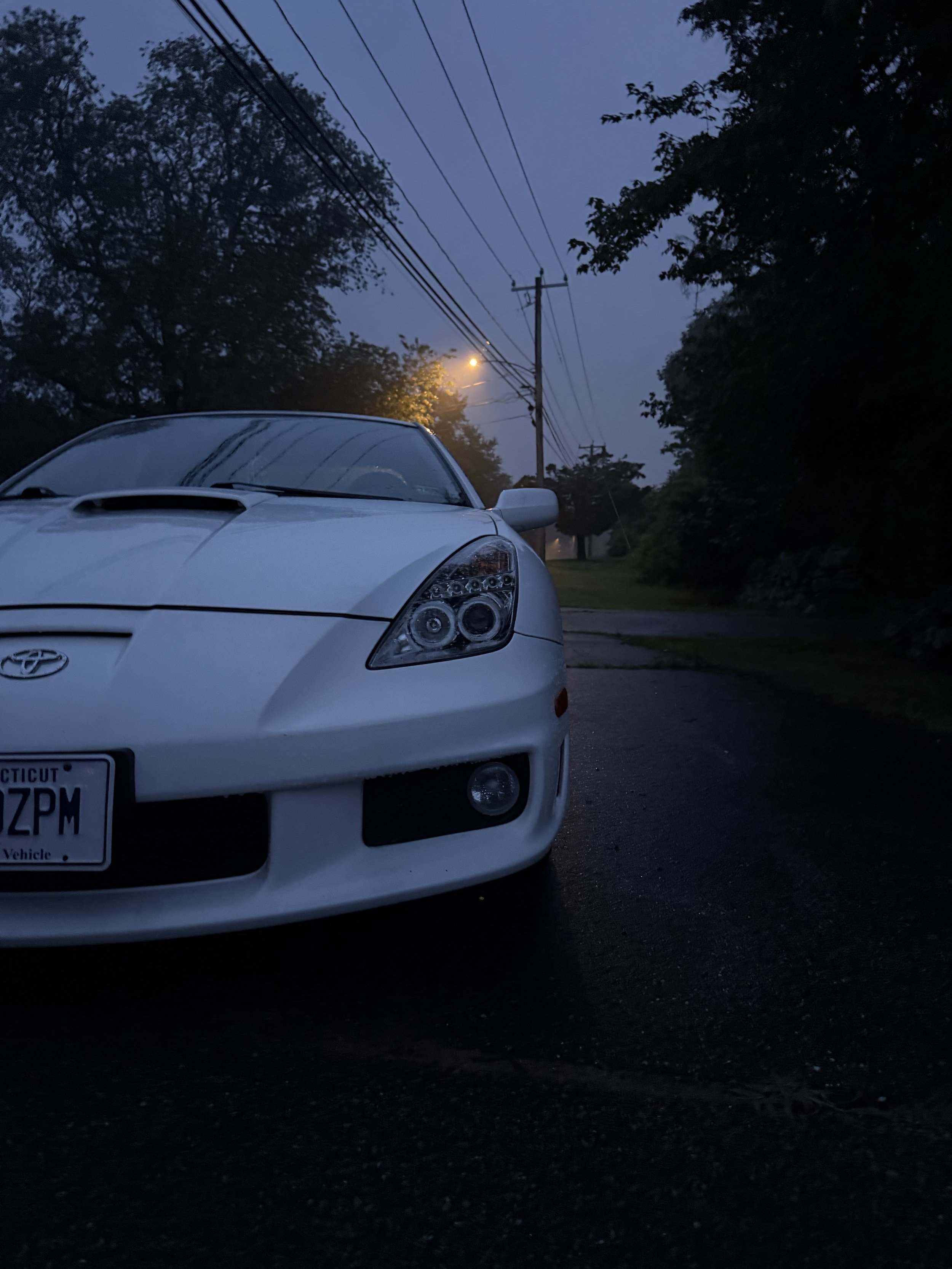 A white car parked on a street during dusk with trees and utility poles in the background.