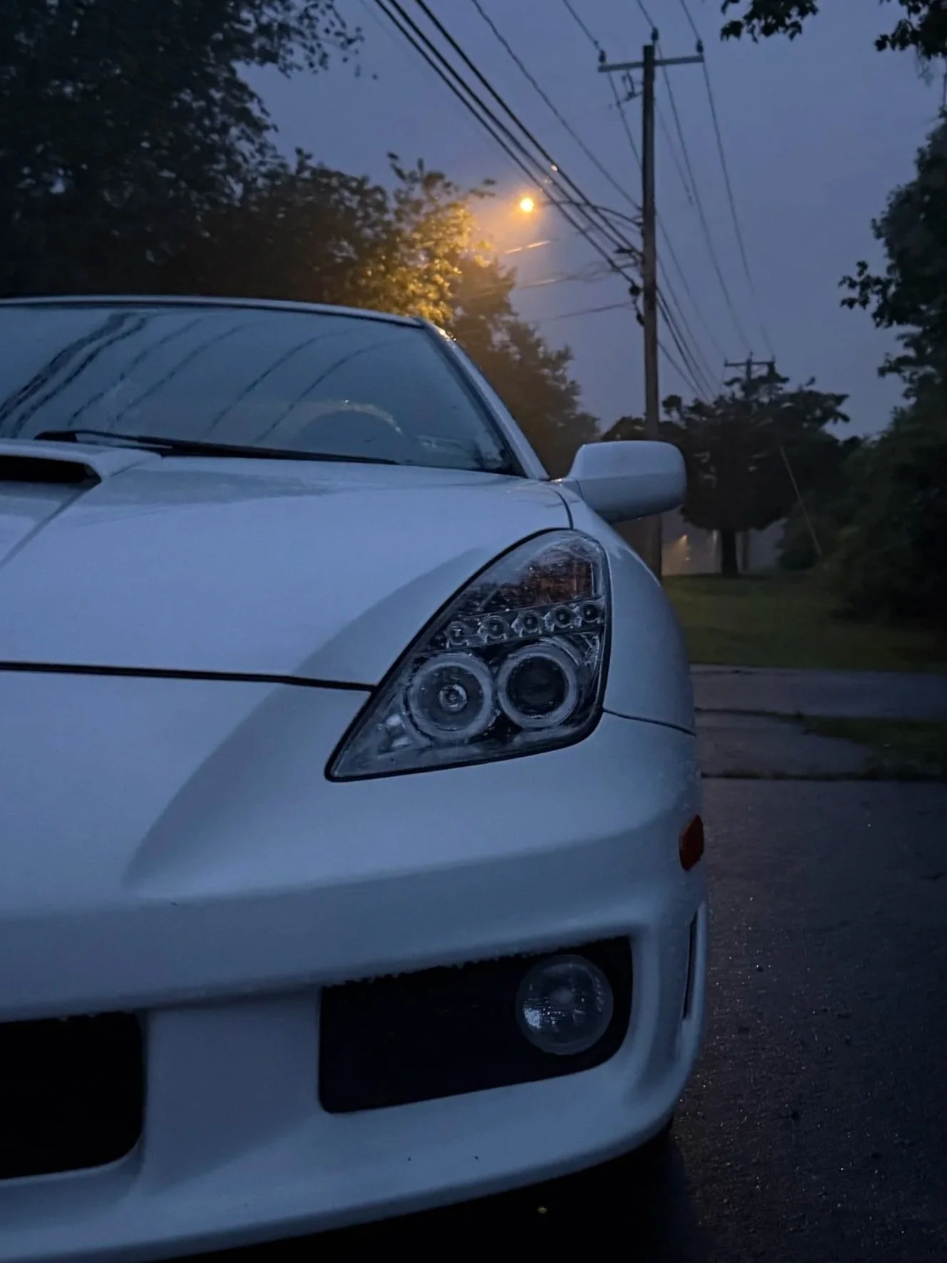 Close-up of a white car parked on a street during dusk or dawn, with trees, power lines, and street lights in the background.