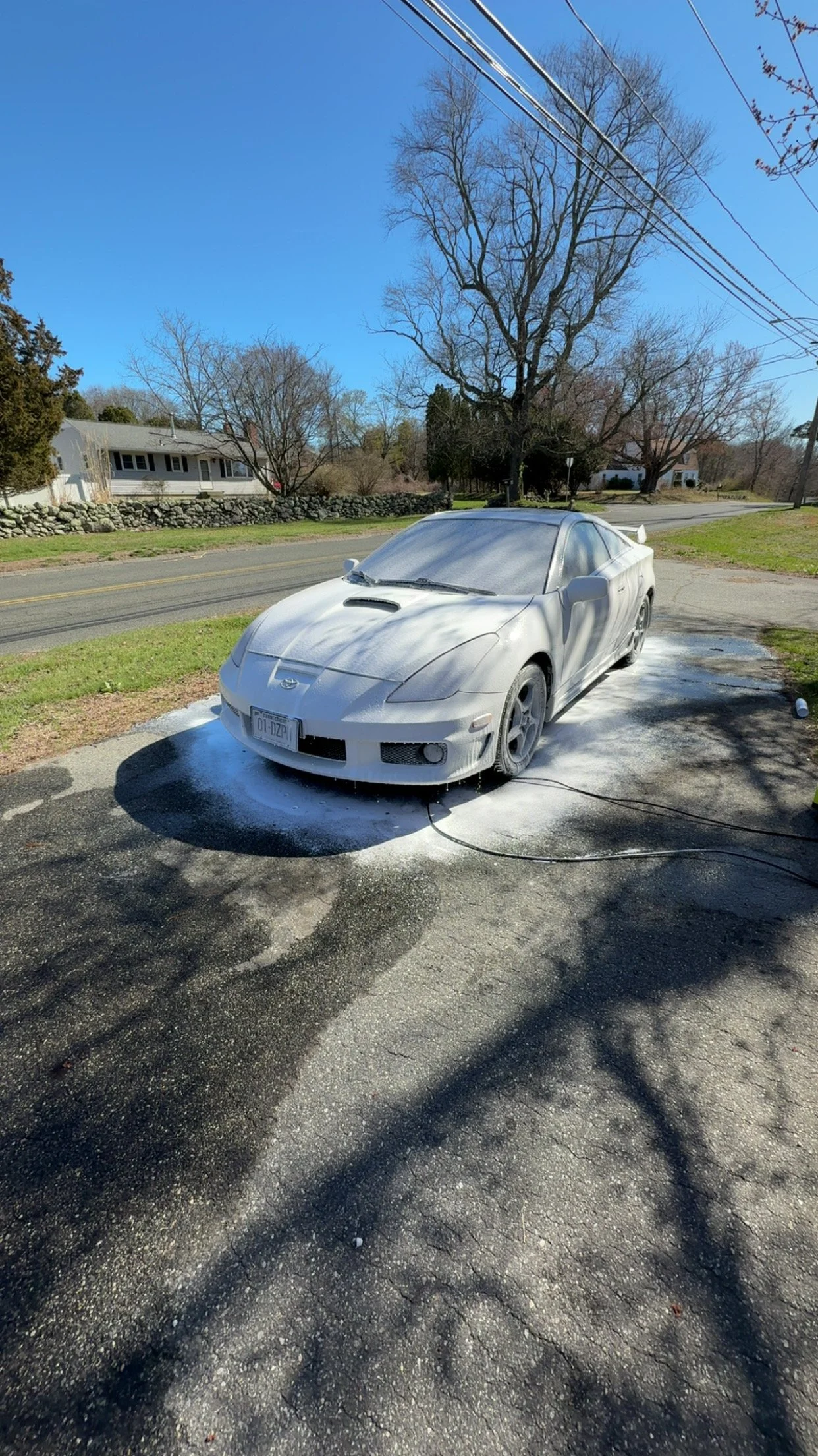 A white sports car covered in soap, parked on the driveway during a sunny day, with foam spilled on the ground around it.