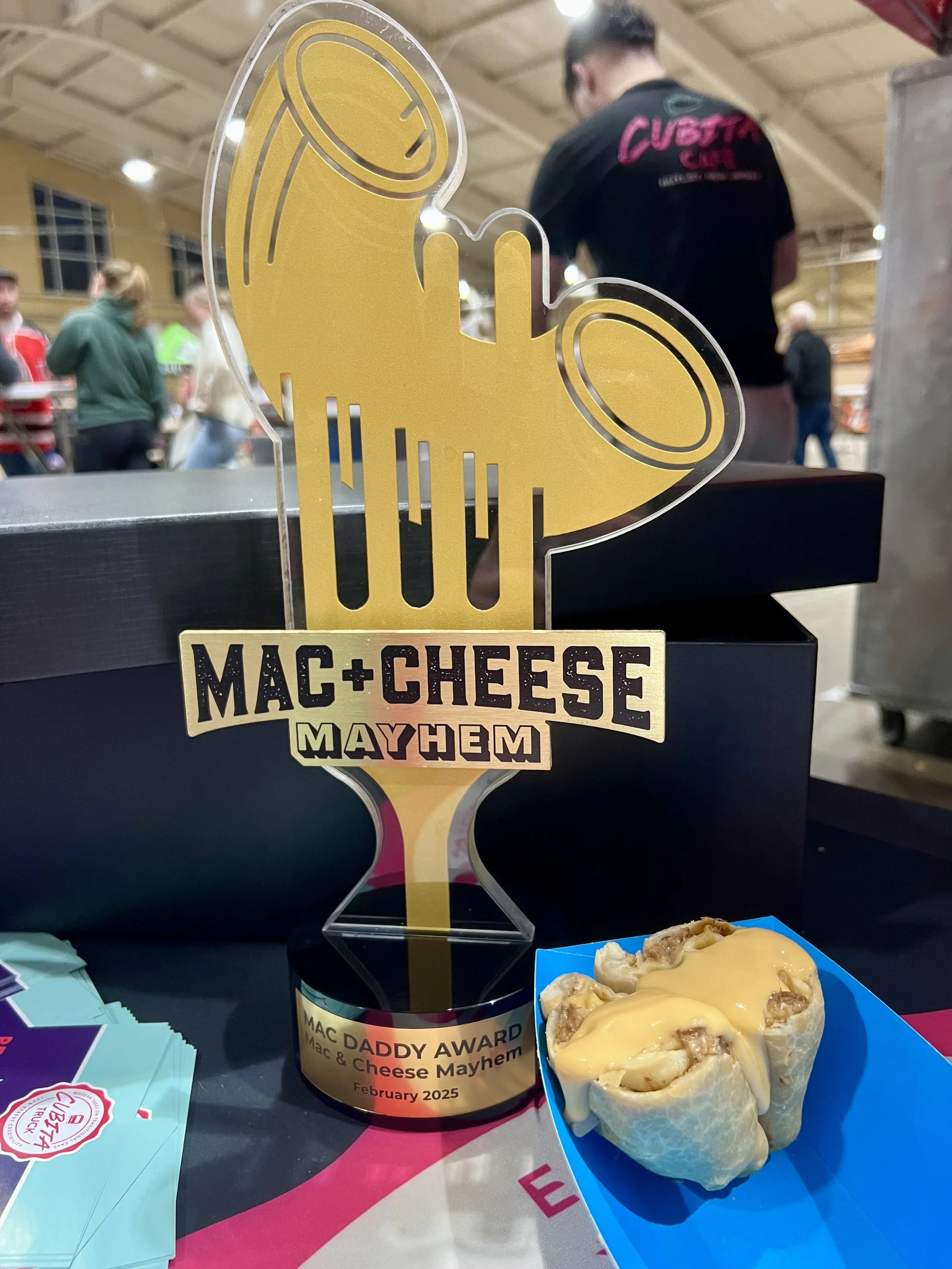 A transparent award trophy with a yellow design of a beer mug, with the text 'MAC + CHEESE MAYHEM' and 'MAC DADDY AWARD' on it. The award is dated February 2025. Next to the trophy, there is a dish of baked macaroni and cheese with cheese sauce and toppings.