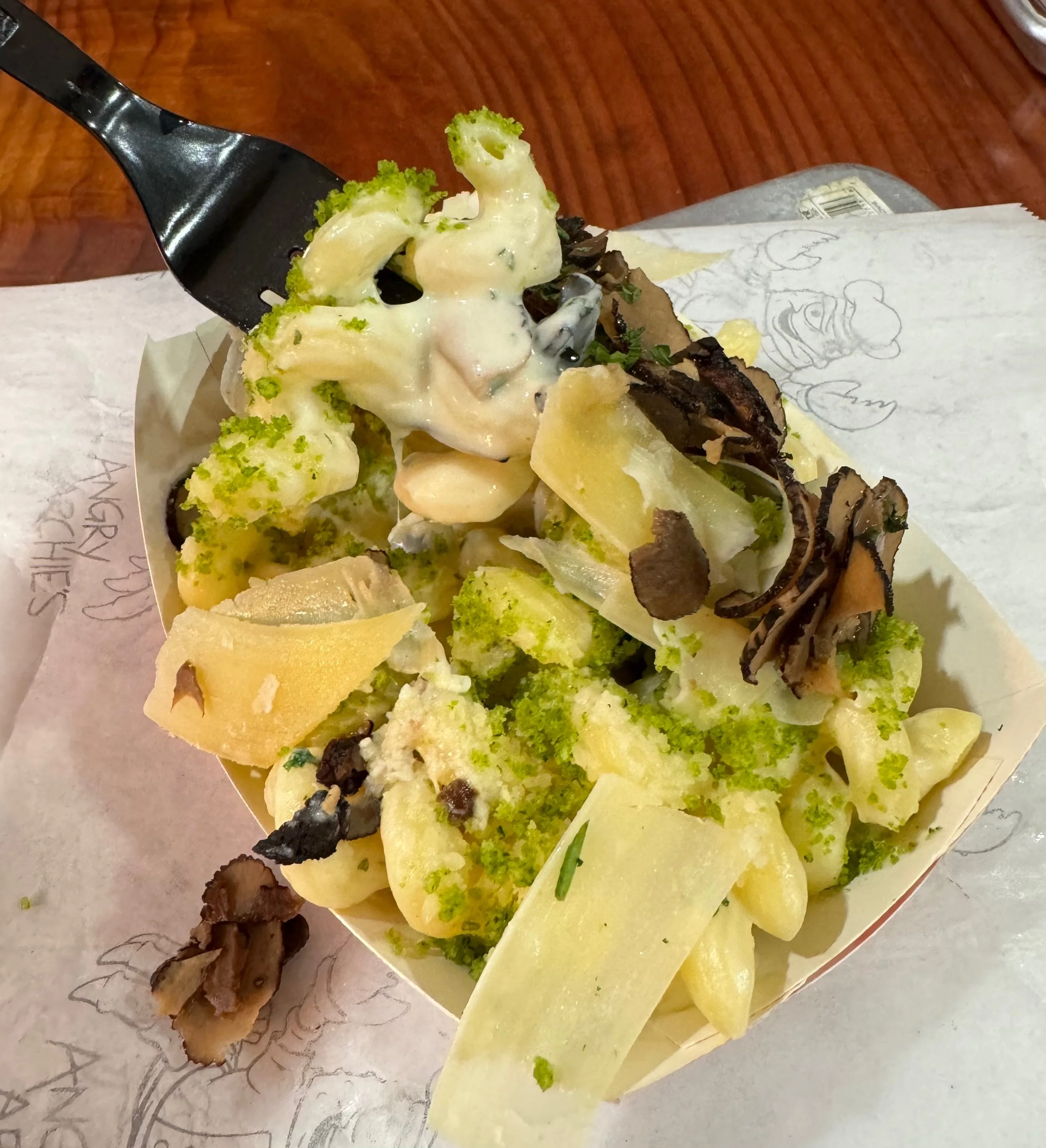 Close-up of a cheese, broccoli, and mushroom pizza with shaved cheese and herbs on top, on parchment paper with a wooden table underneath.