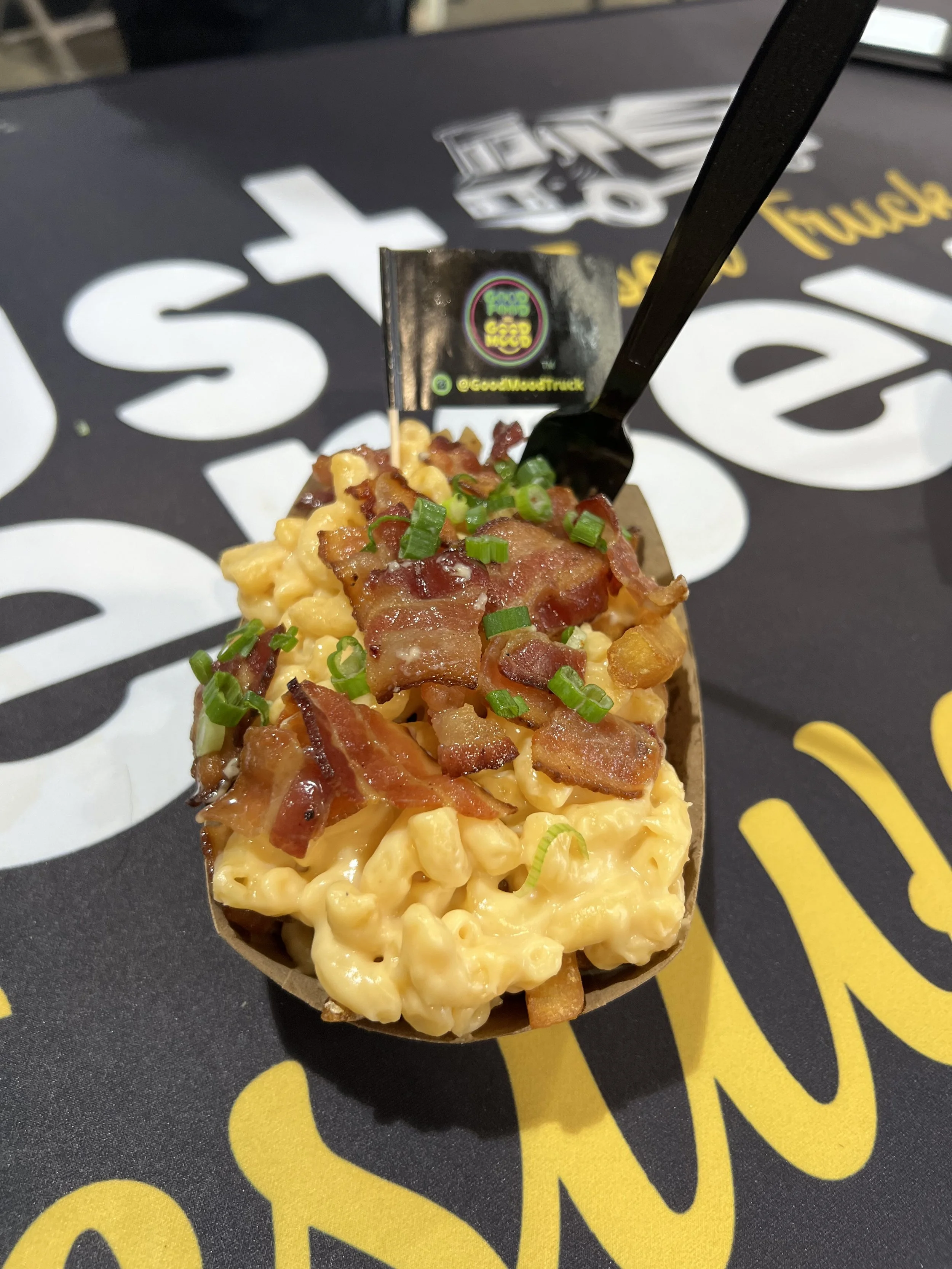 A serving of macaroni and cheese topped with bacon bits and chopped green onions in a paper bowl on a table with a black and yellow logo background.