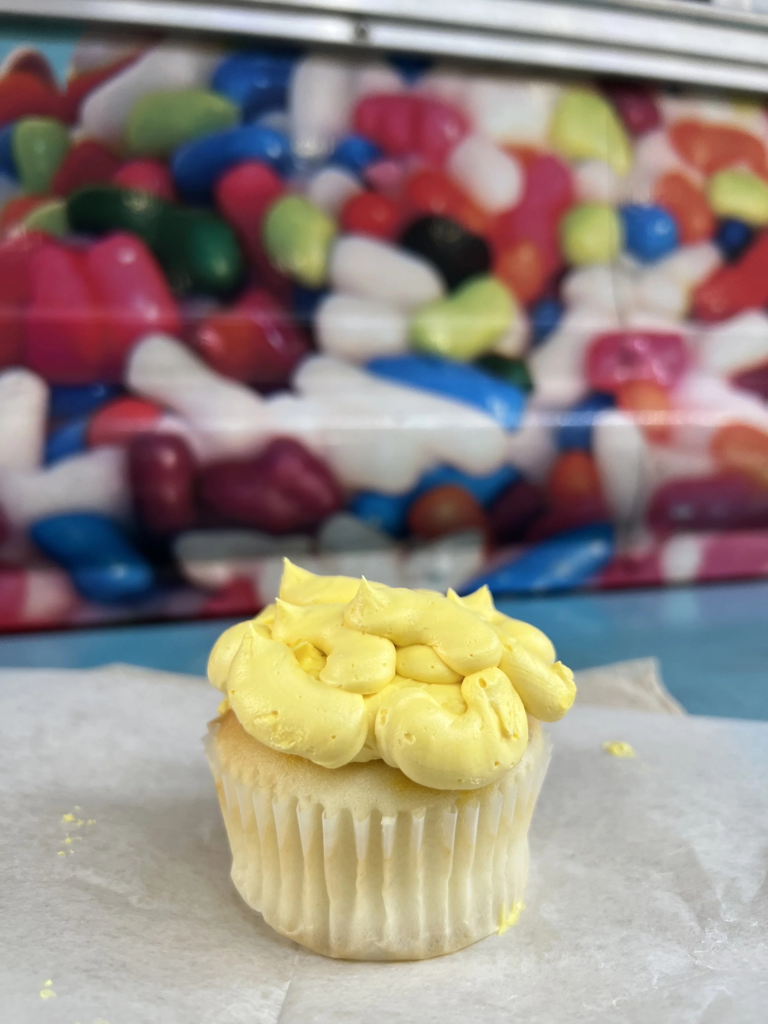 Cupcake with light yellow frosting topped with a cluster of piped yellow frosting flowers, with a blurred background of colorful candies in a clear container.