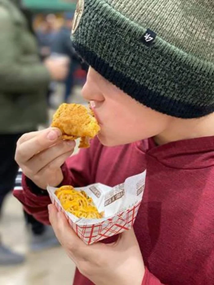 A young person wearing a green and black knit beanie and a red hoodie holding a paper tray with spaghetti and fried chicken, about to eat a piece of fried chicken.