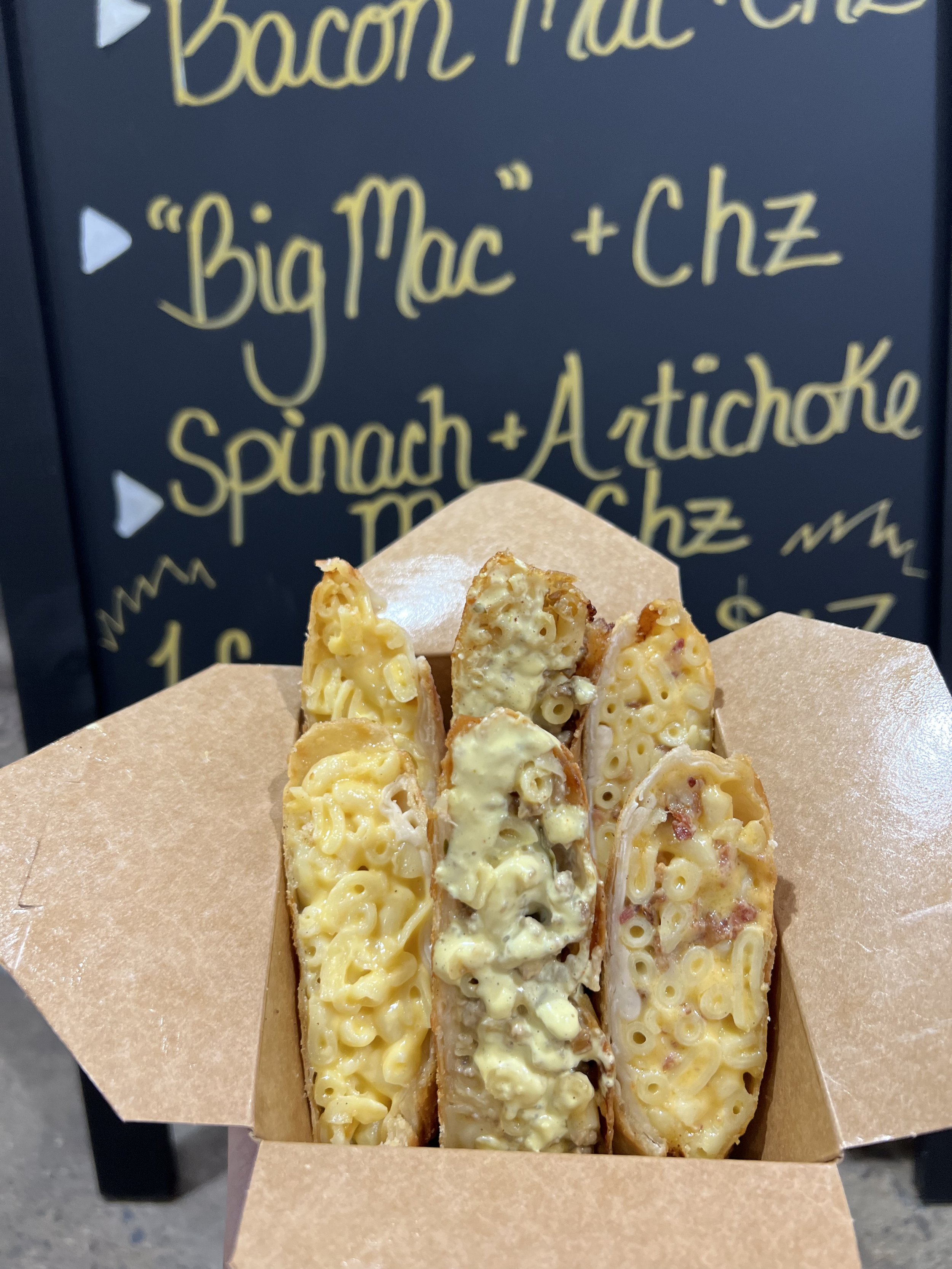 Close-up of three macaroni and cheese stuffed breadsticks in a paper tray, with a chalkboard background featuring a recipe for bacon macaroni and cheese.