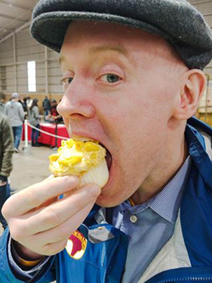 A man wearing a gray cap and blue jacket is eating a cupcake with yellow frosting in a crowded indoor space.