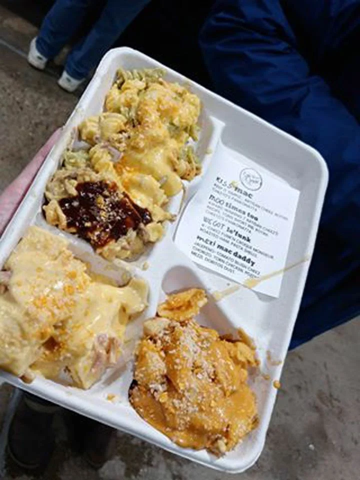 A white tray with four portions of baked macaroni and cheese, some topped with cheese and sauces, a small container with a receipt, and a person's hand holding the tray.