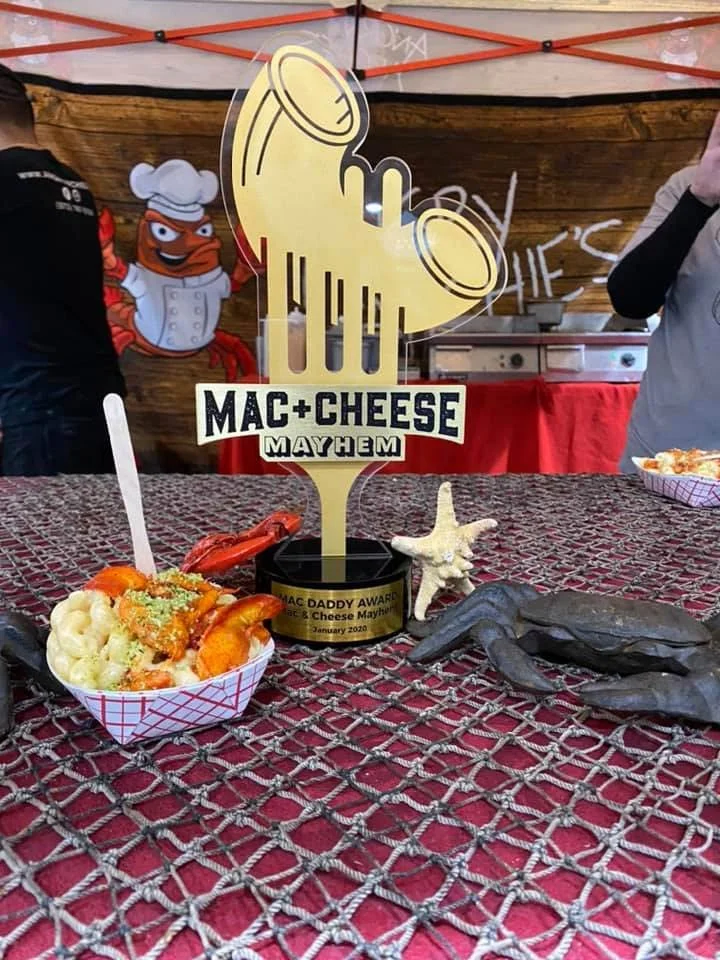 A food tray with macaroni and cheese and lobster, along with assorted shellfish, on a netted table. In the background, there is a large 'Mac + Cheese Mayhem' trophy shaped like a fork with a bow, and a chef mascot poster.