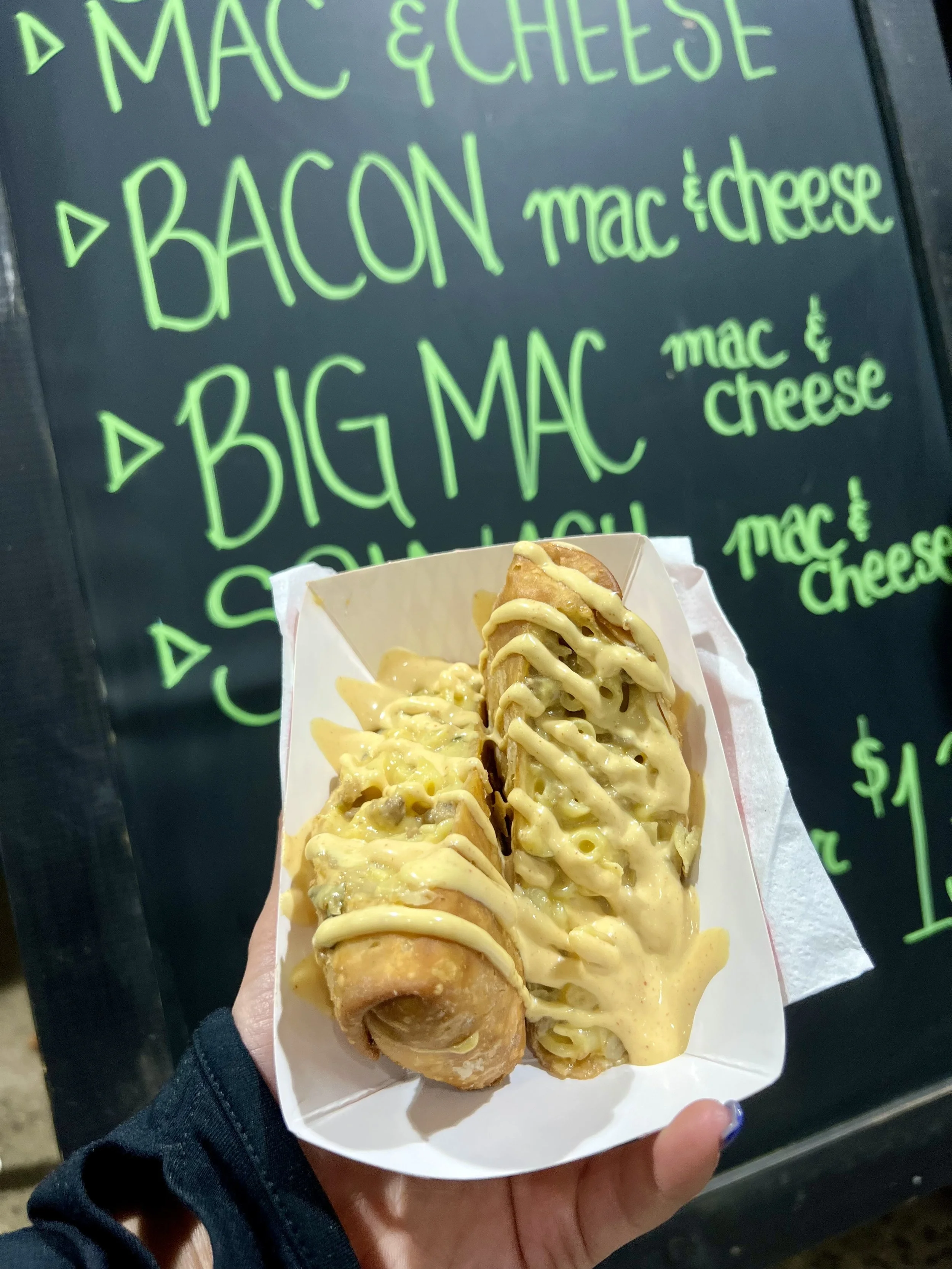 A person holding a white tray with fried spring rolls topped with melted cheese and drizzled with a creamy sauce, in front of a blackboard menu with bright green writing listing various food items.