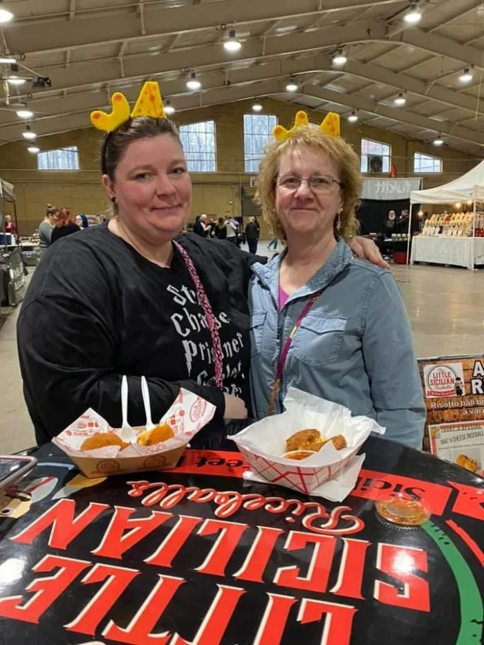 Two women wearing yellow crown headbands with reindeer antlers, standing behind a food stall at an indoor market. The woman on the left has dark hair and is wearing a black shirt, and the woman on the right has light hair and is wearing glasses and a