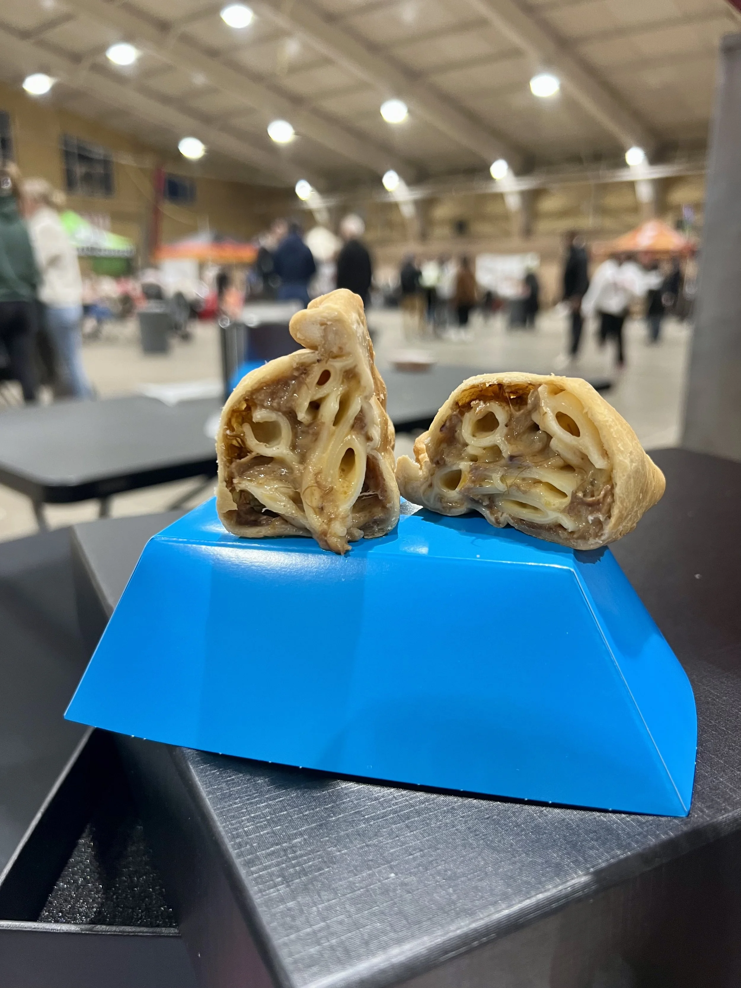 Close-up of two slices of pasta pie with cheese and meat filling on a blue plate in a market hall with blurry people in the background.