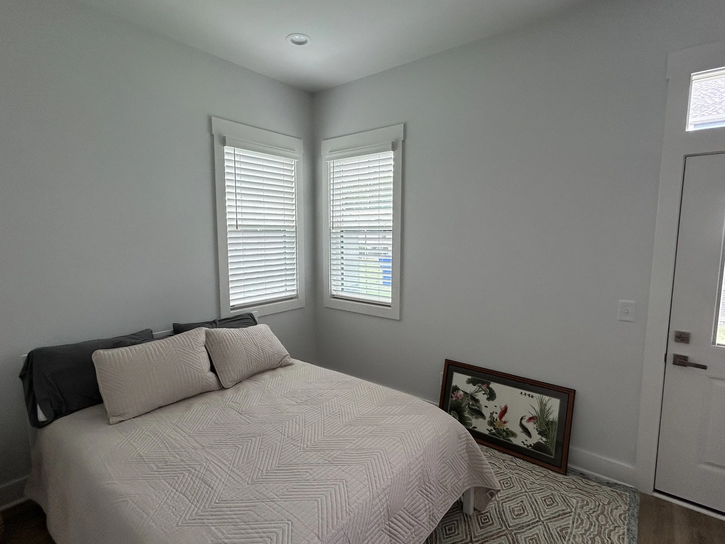 A bedroom with two windows covered by white blinds, a bed with beige quilted bedspread and pillows, and framed artwork of flowers leaning against the wall.