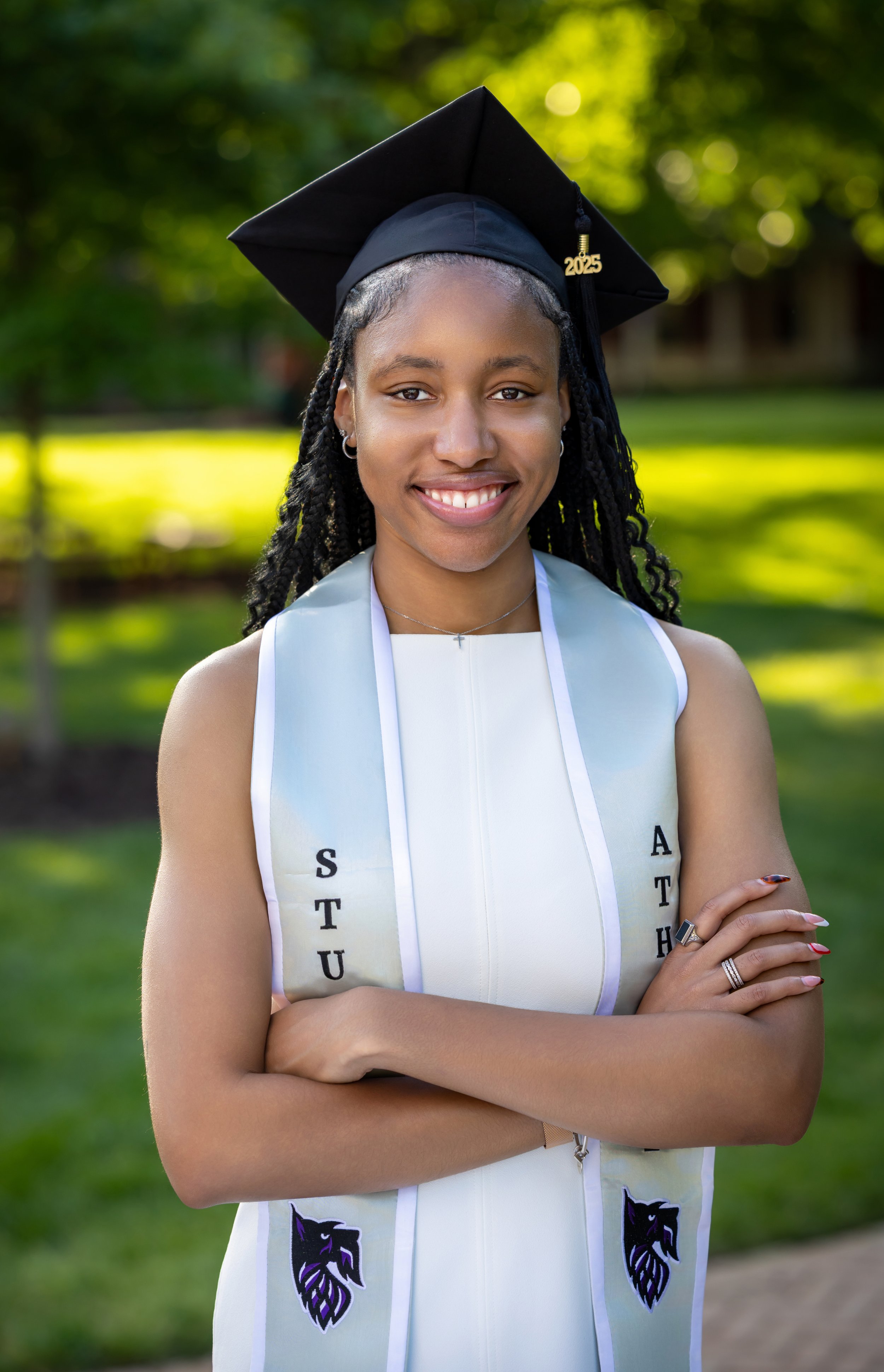 A young woman in a white graduation dress and black cap with a '2025' tassel stands outdoors with her arms crossed, smiling, with trees and sunlight in the background.