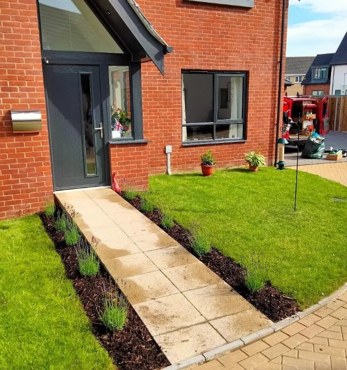 A newly landscaped front yard with a paved walkway, green grass, small plants along the sidewalk, and a brick house with black-framed windows and a door. There are potted plants and gardening tools on the side yard in the background.