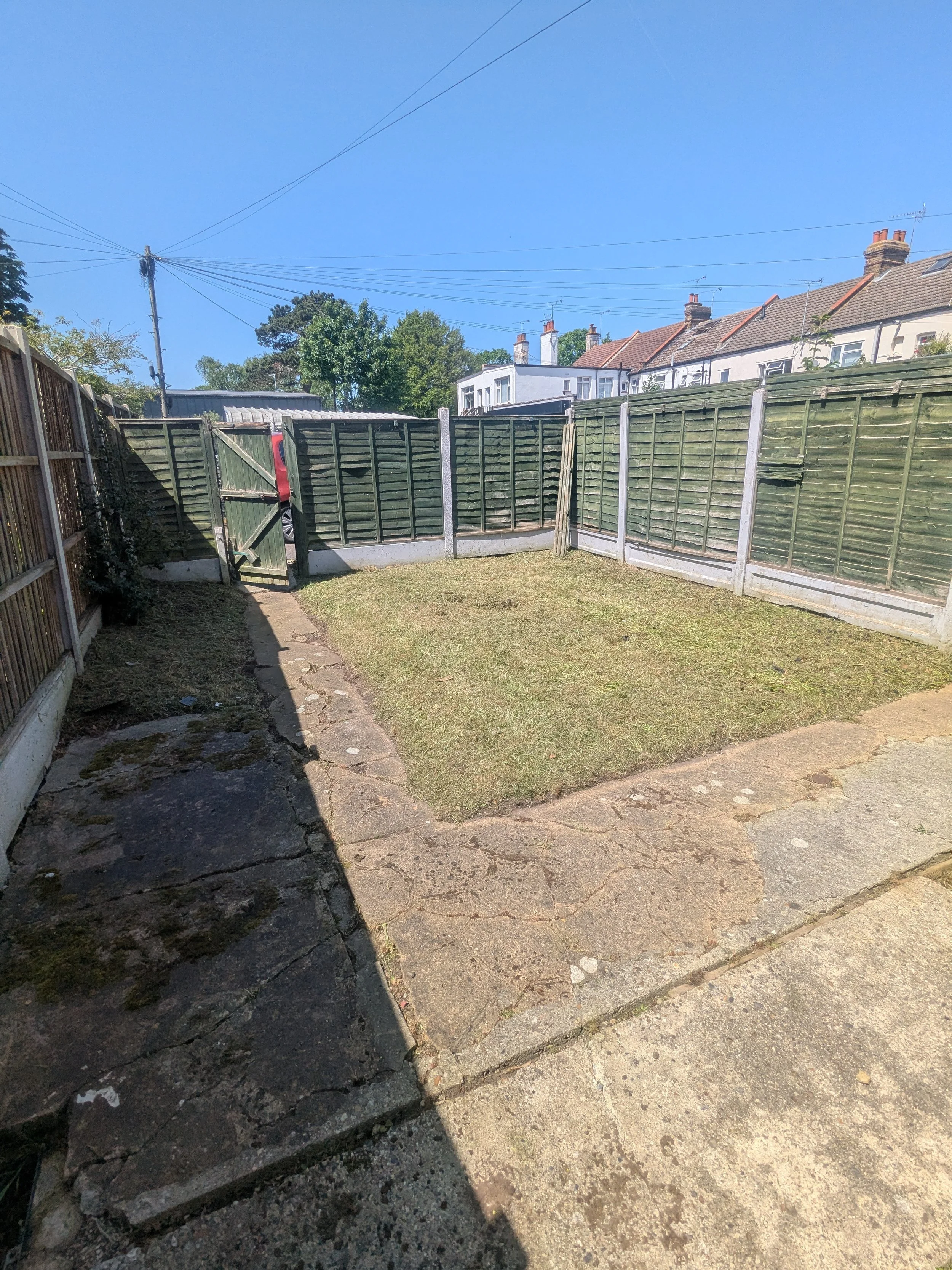 Backyard with a green wooden fence, a small door, a grassy area, and a cracked concrete patio under a clear blue sky. Houses with brick chimneys are visible in the background.
