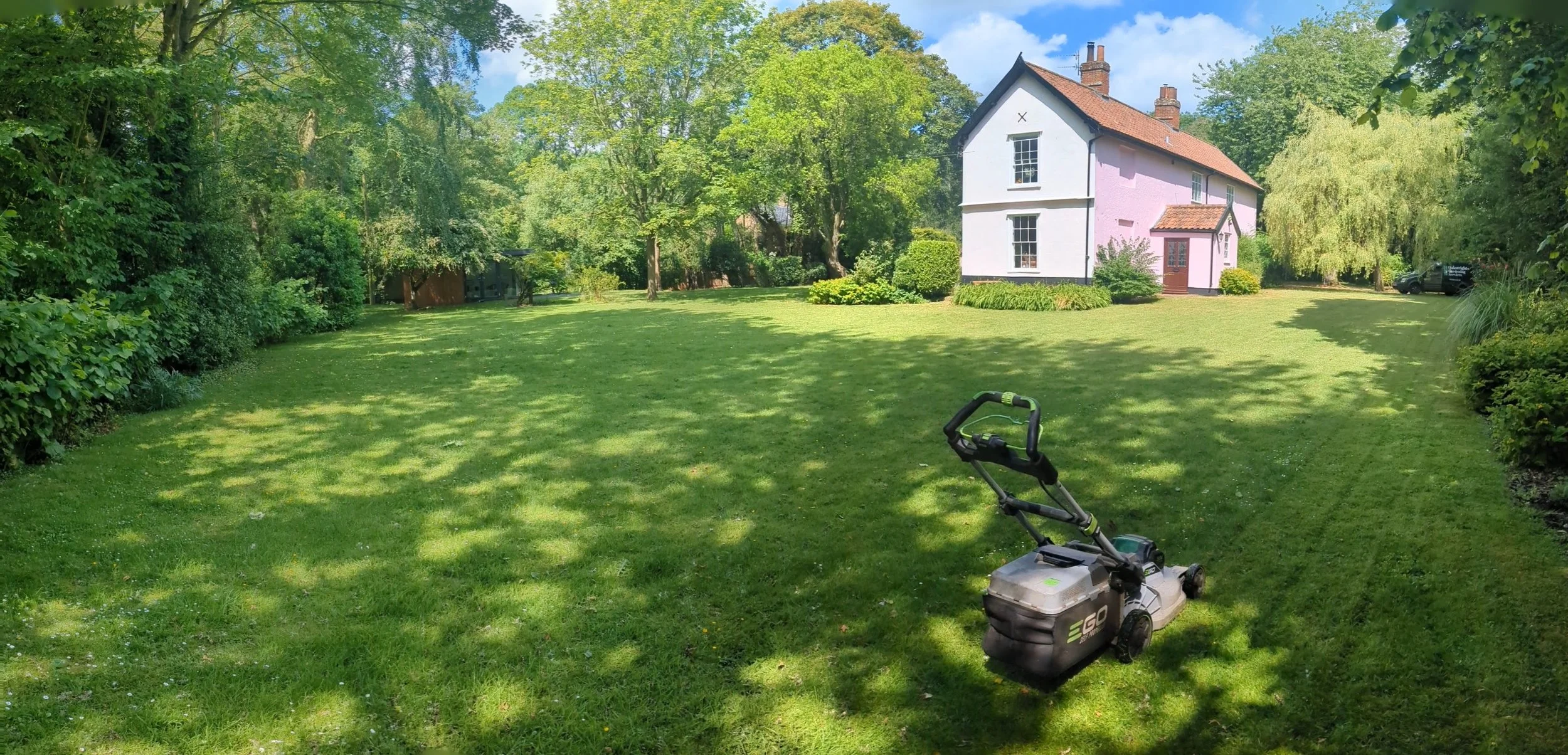 Lush green backyard with a house in the background and a lawnmower in the foreground on a sunny day.