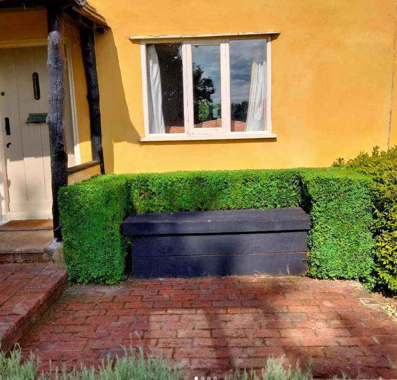 Black wooden bench in front of a yellow house with a window, facing a brick walkway, surrounded by green hedges and bushes.