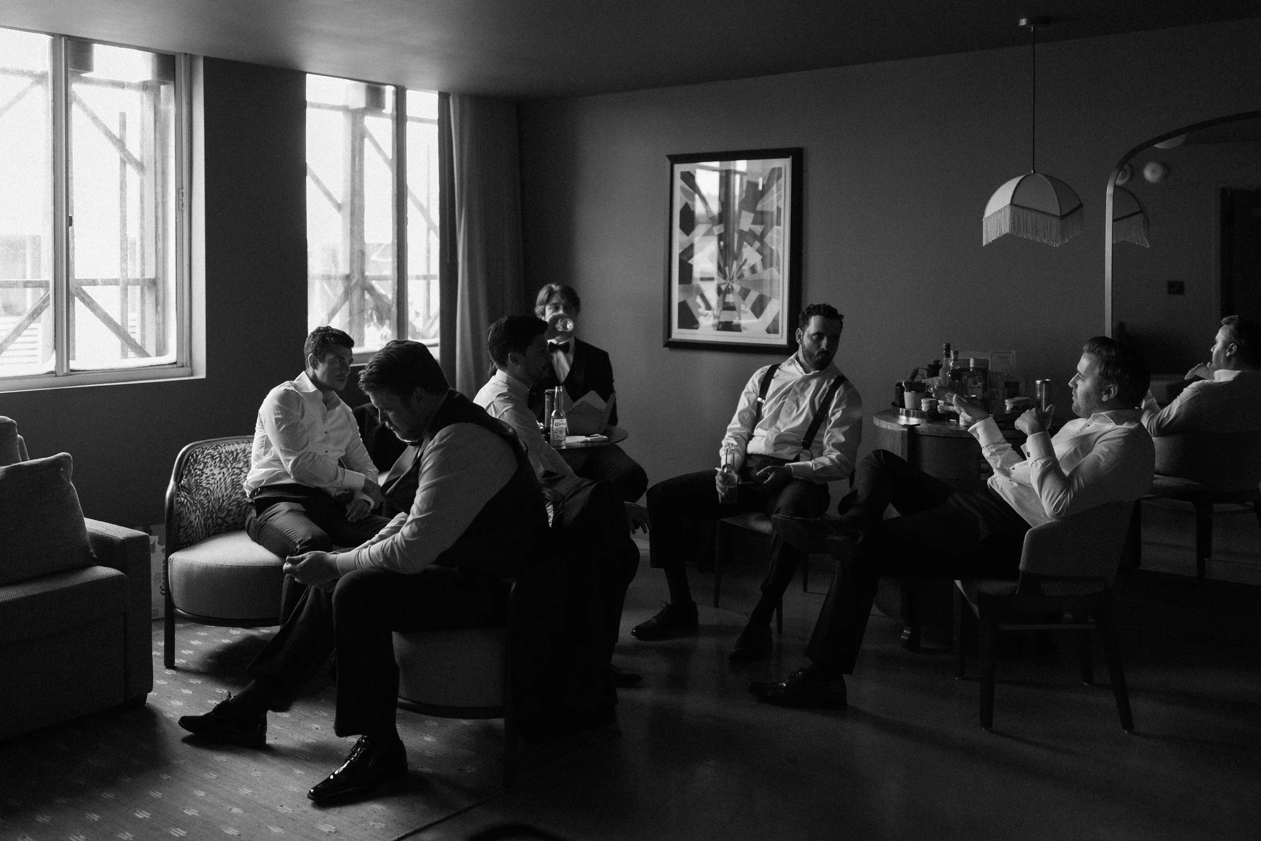 Black and white photo of men in formal attire sitting and standing in a living room. Some are conversing, others are drinking. Large windows with curtains let in natural light. A piece of abstract art hangs on the wall. There is a bar with bottles an