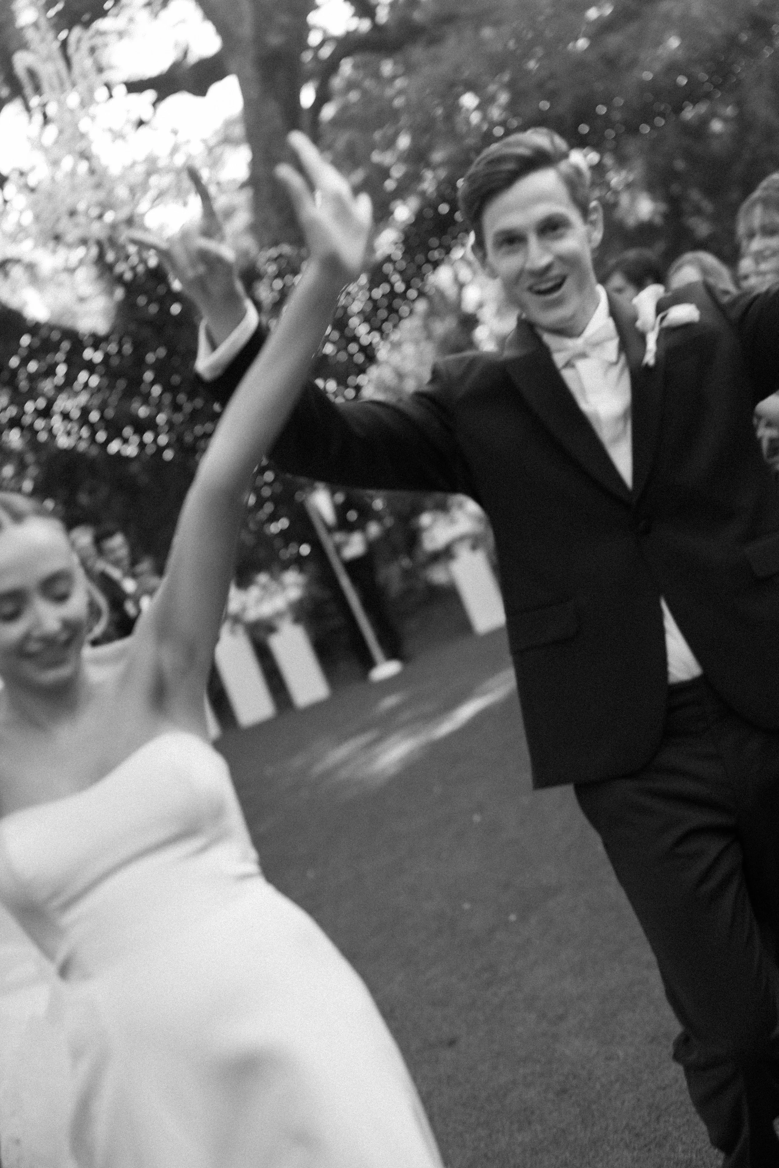 A young man in a tuxedo dancing with a girl in a white dress at an outdoor event, surrounded by trees and other people.
