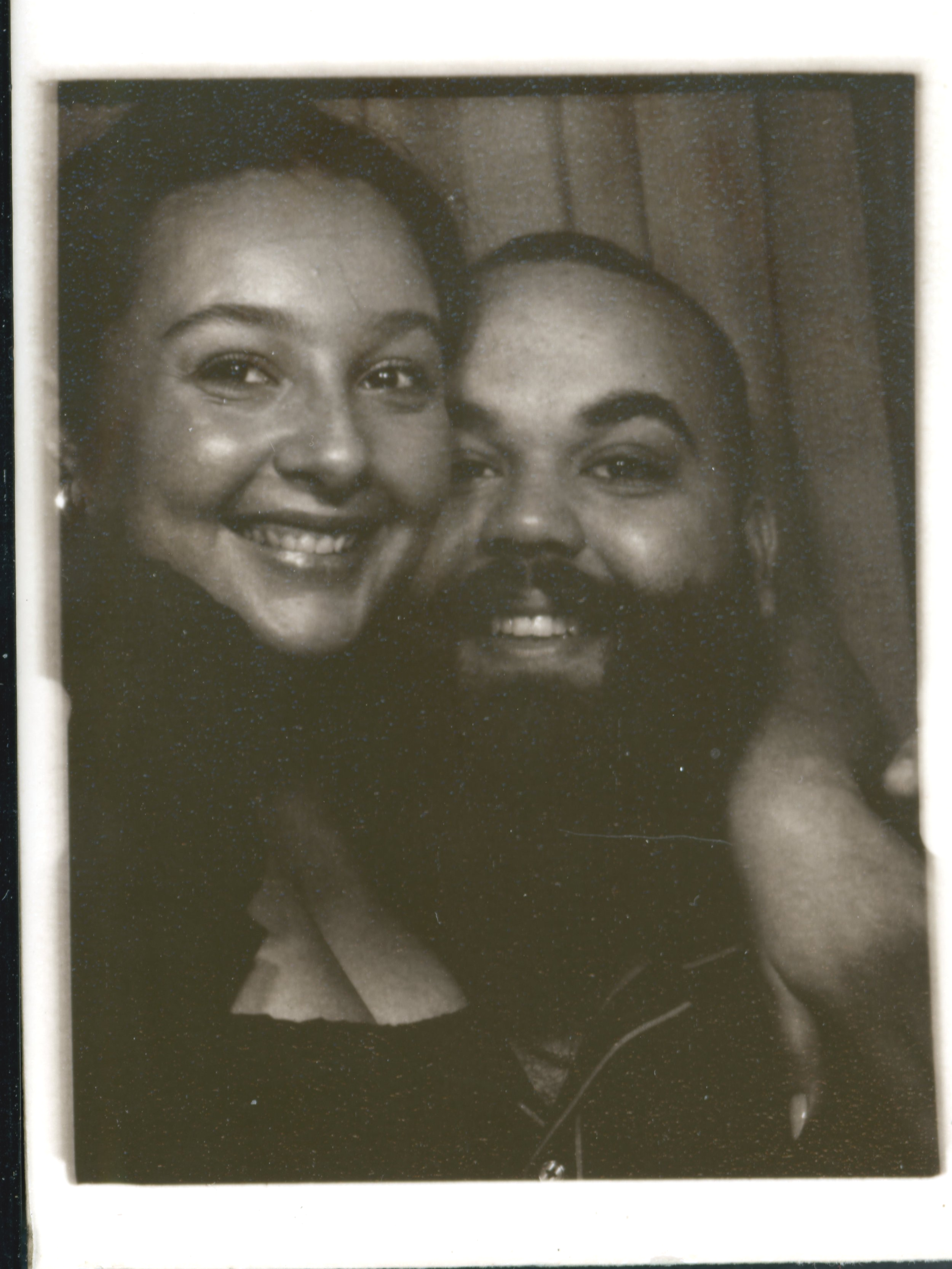 A black and white photograph of two smiling women close together, with one woman on the left and the other on the right. They are looking at the camera, with a wooden ceiling visible in the background.